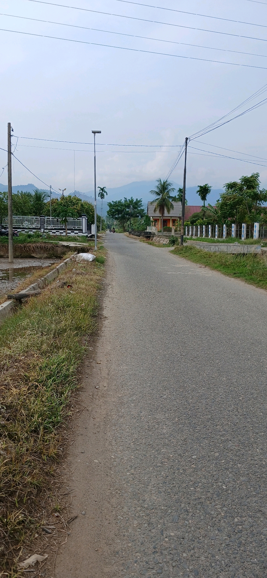 The image shows a street in Glumpangpayong, Indonesia. The road is paved and appears to be a two-lane street. On the left side of the road, there is a drainage ditch with overgrown grass and some debris. Beyond the ditch, there is a white fence with black accents, and behind it, trees and some buildings. On the right side of the road, there are more trees, houses, and a decorative fence with blue and white posts. Power lines are visible overhead, supported by utility poles. In the distance, mountains can be seen under a cloudy sky. A single street light is visible on the left side of the road. A motorcycle is seen in the distance on the road. The overall scene depicts a rural or semi-rural residential area.