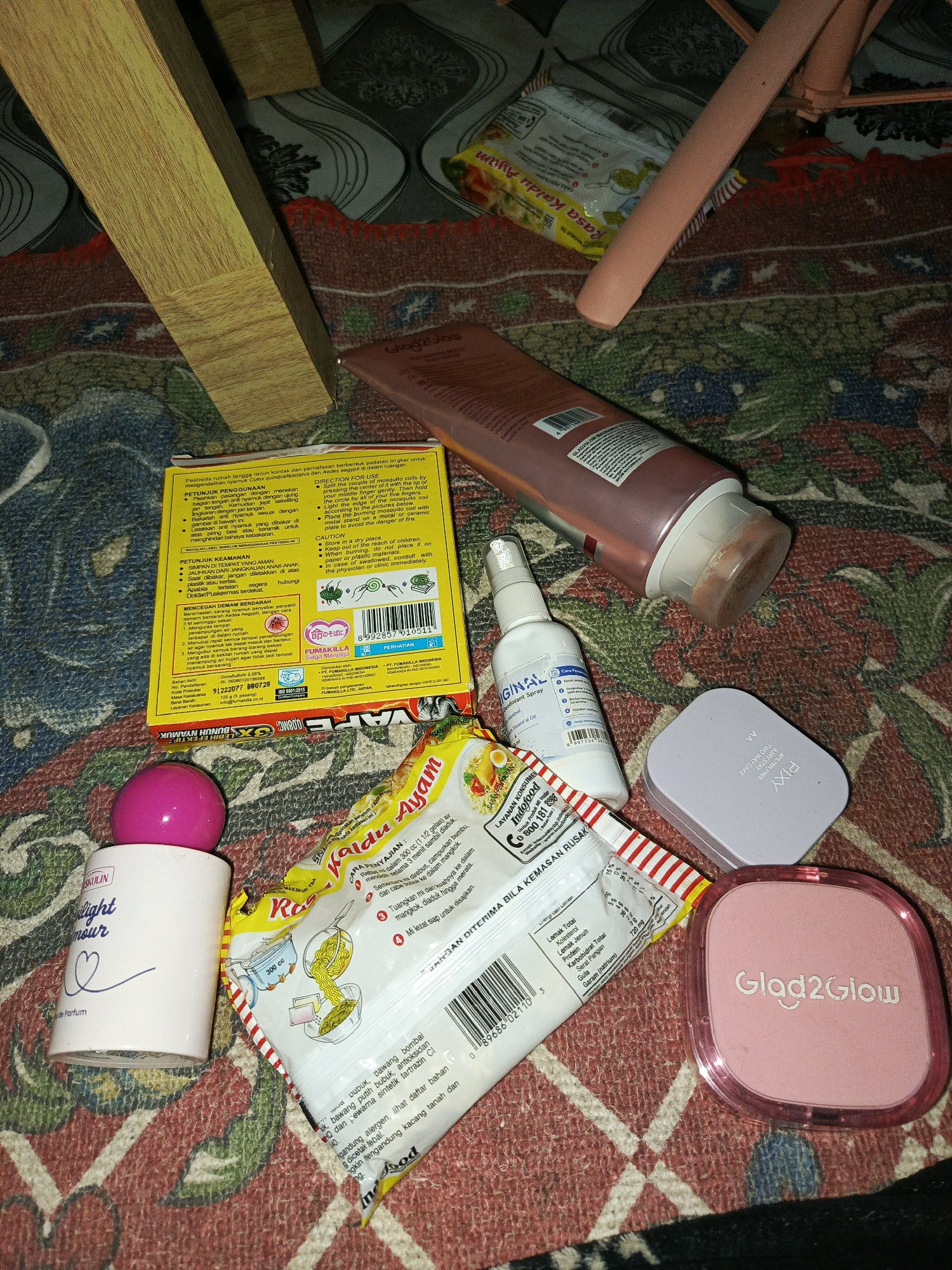 A close-up, overhead shot shows a collection of personal care items and food packaging arranged on a patterned rug. Among the items are a pink bottle with a white label and pink spherical cap that reads "SKULIN light amour de Parfum", a white spray bottle labeled "ORIGINAL Deodorant Spray", a pink compact with the brand "Glad2Glow" on it, and a light purple compact with "PIXY" printed on it. There is also a yellow box with Indonesian text, and a packet of instant noodles with an illustration of noodles being prepared. The scene appears to be indoors, possibly in a home, on a rug with a red, black, and blue pattern. The lighting is dim, suggesting it might be nighttime or a poorly lit room. There is no indication of the city of Megang Sakti I, Indonesia.