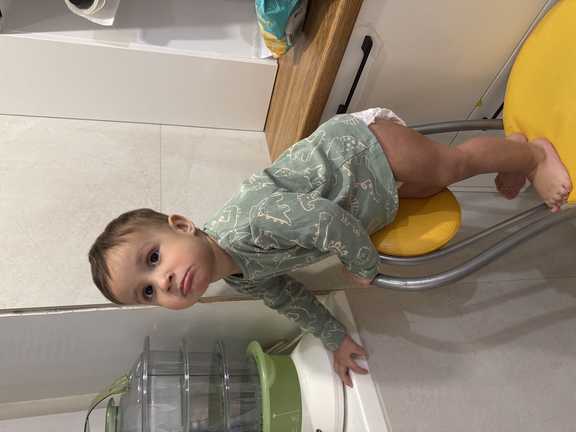 A young child wearing a light green dinosaur-patterned pajama top and a diaper is perched on a yellow padded stool with a silver frame. The child leans towards a white countertop where a green and white food steamer sits. The child is positioned near a white cabinet and a wooden shelf or countertop is visible in the upper right. The setting appears to be indoors, possibly a kitchen. There is no information provided about the location being in the city of Chaikovsky, Russia.