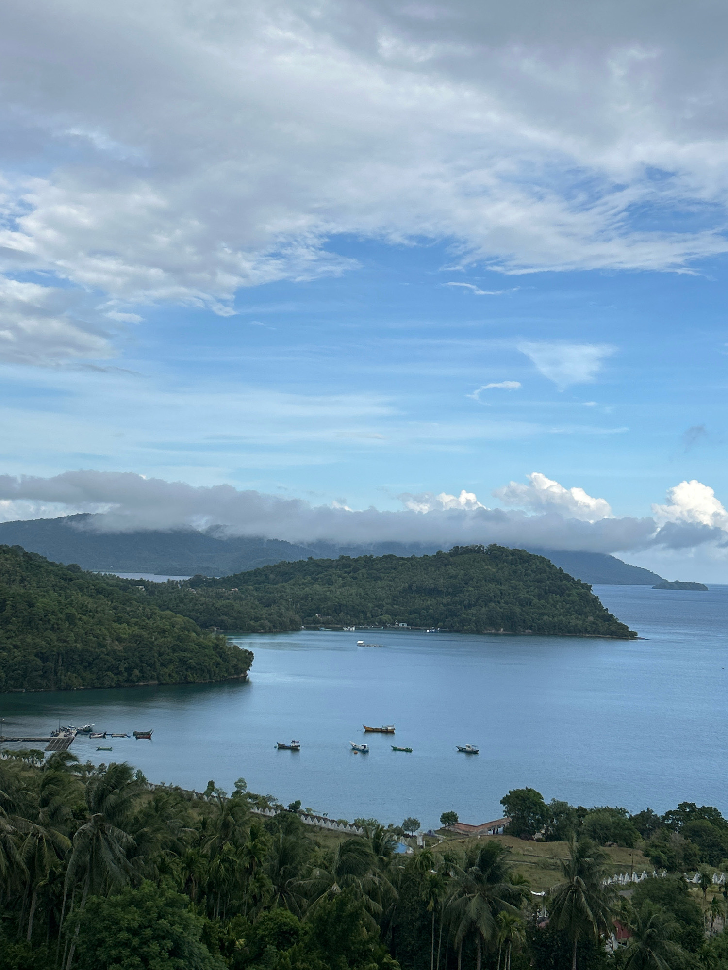 This image captures a scenic view of the coastline near Sabang, Indonesia. In the foreground, lush green palm trees and other vegetation line a slightly sloped area with a white, low-lying wall running horizontally. Beyond this, a calm, blue body of water dominates the midground, dotted with several small boats. The water is bordered by verdant, tree-covered hills that rise gently from the shore. In the background, larger, mountainous islands stretch across the horizon, some topped with wispy clouds that partially obscure their peaks. The sky above is a blend of blue sky and white, textured clouds, suggesting a partly cloudy day. The overall scene conveys a serene and natural coastal landscape.