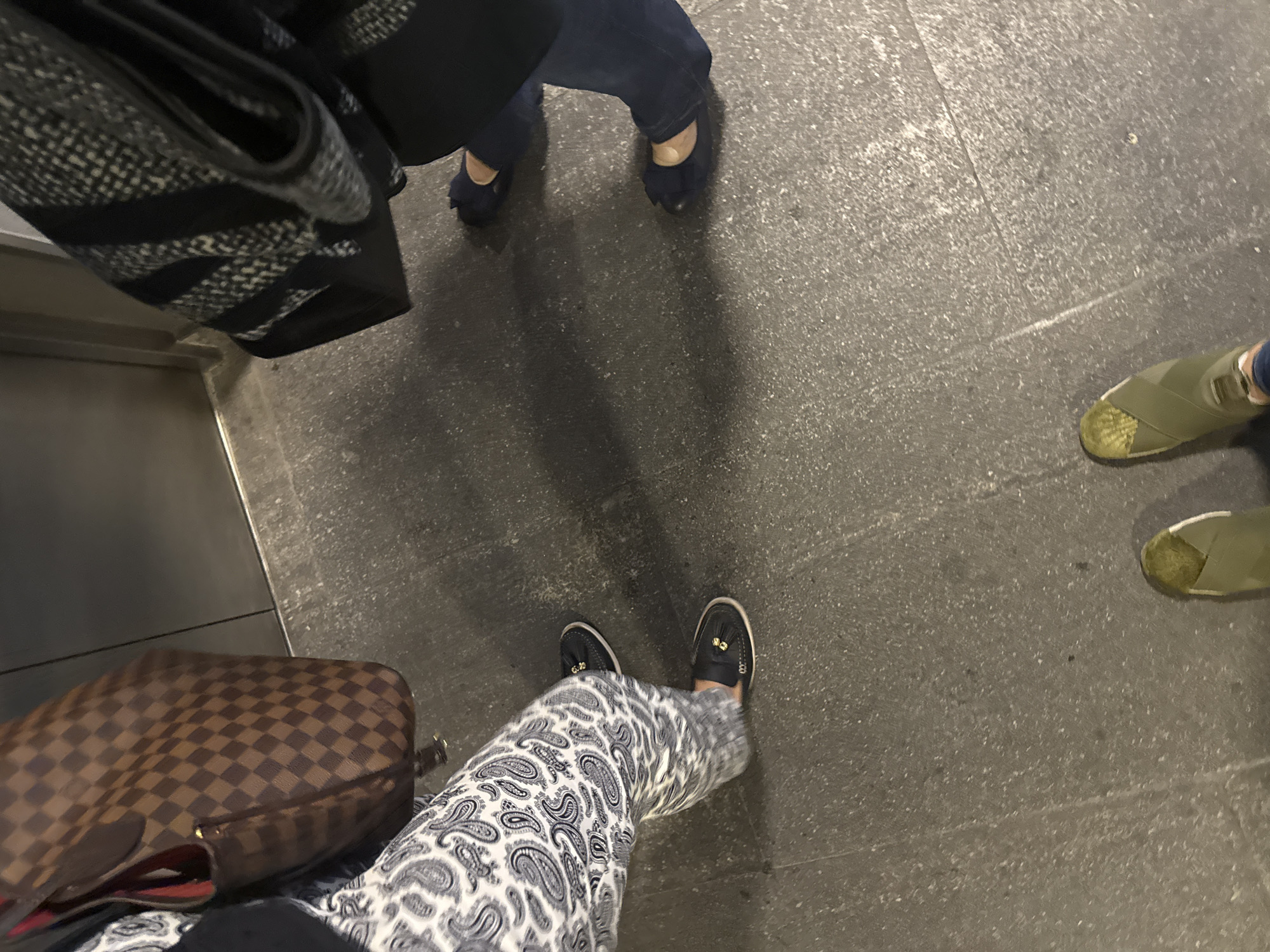 The image displays an overhead-angle, ground-level view of the lower extremities of three individuals standing on a dark gray, speckled floor. In the foreground, a person wears olive green slip-on shoes with a white sole stripe and dark blue jeans. To the left, a second individual is visible from the waist down, wearing white pants with a black paisley pattern and black slip-on shoes featuring gold-colored embellishments. This person also carries a brown handbag with a checkerboard pattern and visible red interior lining. To the right, a third individual's lower legs are shown, clad in dark blue pants and dark blue flat shoes with a bow detail on the toe. This person carries a dark-colored bag with a textured, patterned surface, partially visible. A dark, vertical background element, possibly a wall or door, with a narrow lighter-colored horizontal strip at its base, indicates an enclosed interior space. The scene is located in the city of San Bernardino Tlaxcalancingo, Mexico.