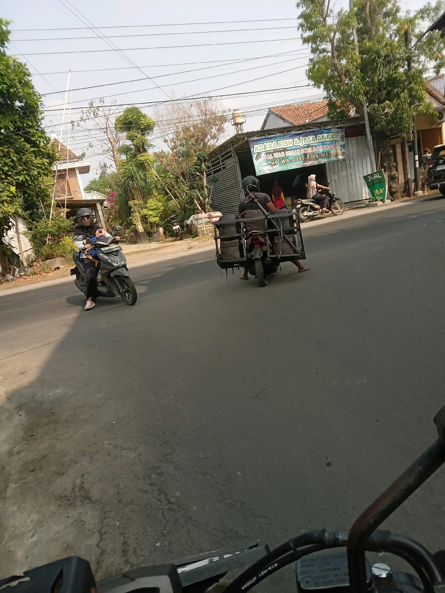 In Banguntapan, Indonesia, on a paved road with a slight incline, a motorcyclist wearing a helmet and hijab rides a dark gray scooter. Behind her, a three-wheeled motorcycle cart is being ridden by two individuals, one appearing to be the driver and the other a passenger. The cart is loaded with several boxes. To the right of the road, a building with a banner that reads "ADARA FISH & JASA ANGKUT" is visible, suggesting a business related to fish and transportation services. The environment is sunny, with trees and overhead power lines present.