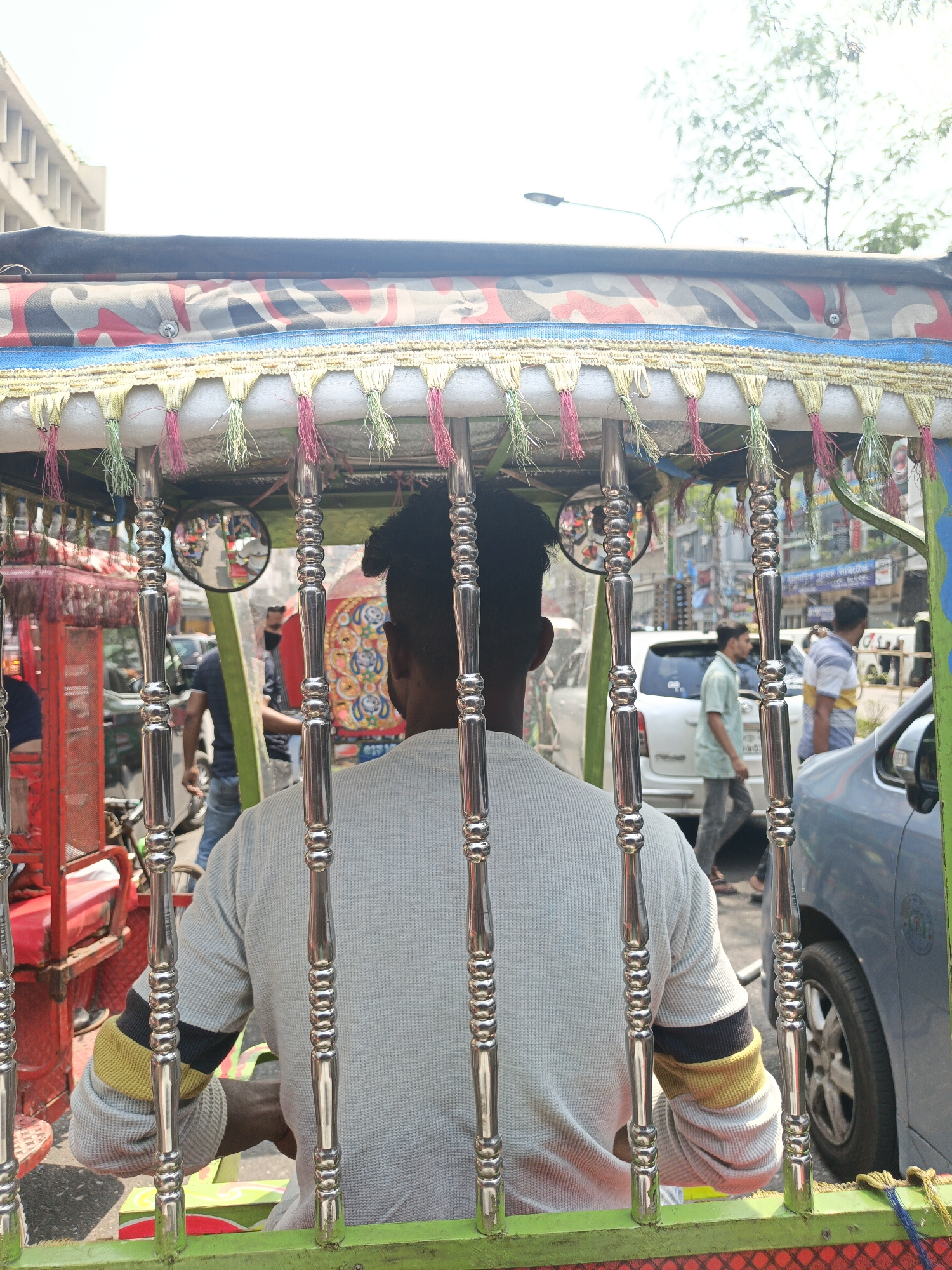 The image shows the back of a rickshaw driver in Dhaka, Bangladesh. The rickshaw has a decorative canopy with tassels and ornate metal bars. The driver is wearing a gray sweater with striped cuffs. In the background, there is a busy street scene with pedestrians, cars, and buildings. The time of day appears to be daytime, with sunlight present.