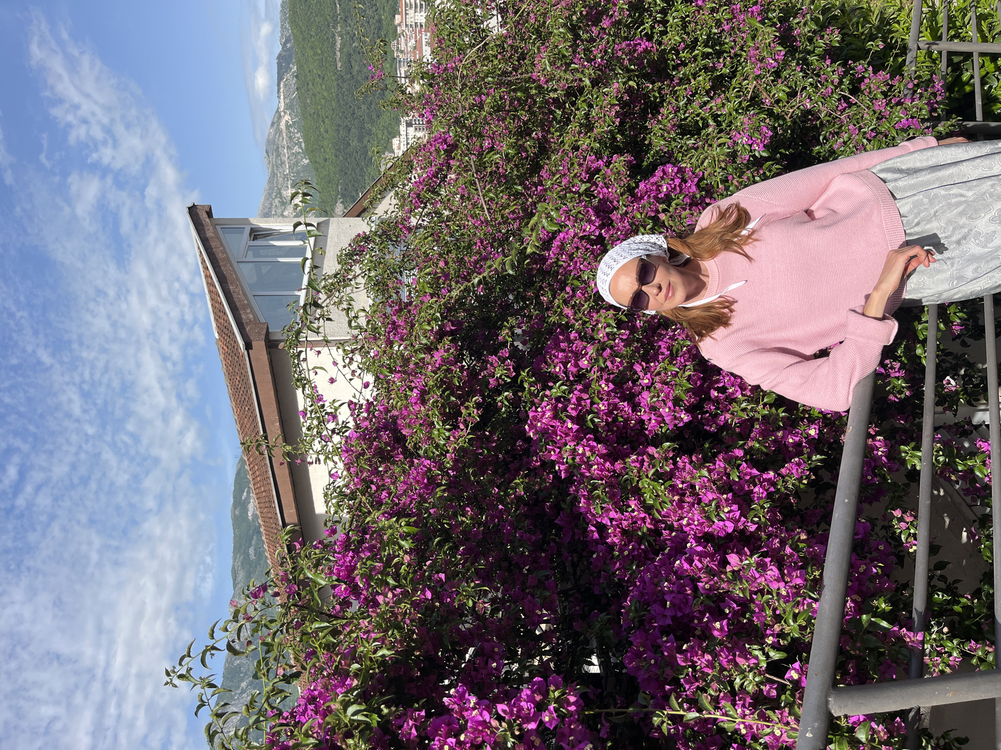 A medium shot shows a light-skinned woman standing next to a flourishing bougainvillea bush in Budva, Montenegro. The woman has brown hair and is wearing sunglasses, a white headscarf, a pink sweater, and a silvery skirt. Her right hand rests on a dark gray metal railing. The bougainvillea is full of vibrant purple blossoms and green leaves.
In the background, there is a residential building with a tan roof. Further back, a scenic landscape with green mountains and a blue sky dotted with clouds is visible. Buildings that could be part of the city of Budva appear at the top of the mountains. The image seems to be taken during daylight, judging by the clear sky and lighting.