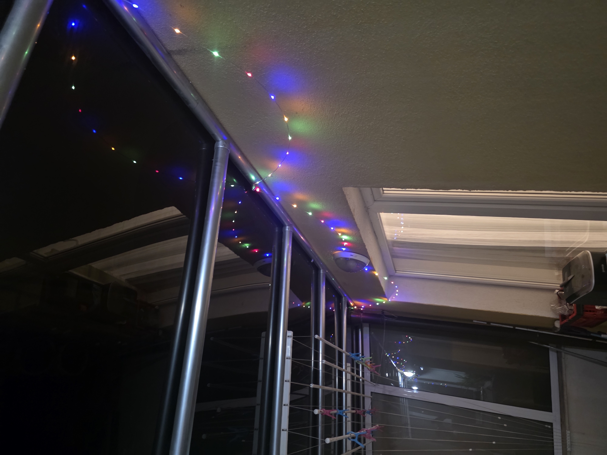 This is a photograph of an outdoor balcony at night, adorned with a string of multi-colored LED lights. The lights are draped along the edge of the ceiling and down the side wall, casting a festive glow. Below the lights, a clothesline with several colorful clothespins is visible. The background features a window with sheer curtains and a portion of a building wall. The overall impression is one of a decorated outdoor space at dusk or night. There is no indication of a specific location within the city of Sofia, Bulgaria.