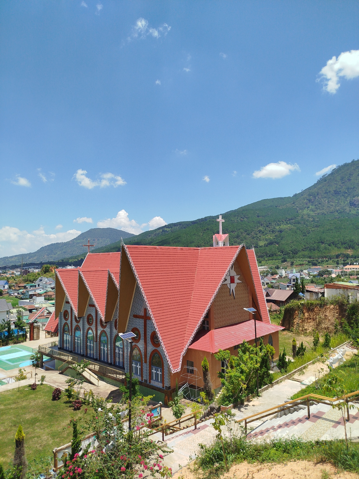 A wide shot captures a church with a red tiled roof and arched windows, likely in Định An, Vietnam, under a bright blue sky dotted with fluffy white clouds. The church's facade is made of grey stone with subtle red cross accents and features a prominent red-tiled roof with decorative white trim. A white cross atop a pink structure stands on the roof. The church is situated on a grassy hill overlooking a town with various buildings and lush green mountains in the background. Several solar panels are visible on the church roof, and pathways with railings lead down the hillside. A swimming pool and some trees are also present in the foreground.