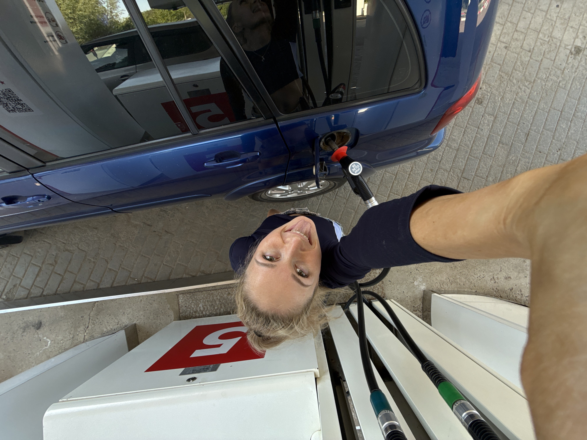 The image shows a woman taking a selfie at a gas station. She's holding the gas pump nozzle inserted into her blue car. The gas pump is labeled with a large red "5". The reflection in the car window shows another person standing nearby. The location is Chaikovsky, Russia.