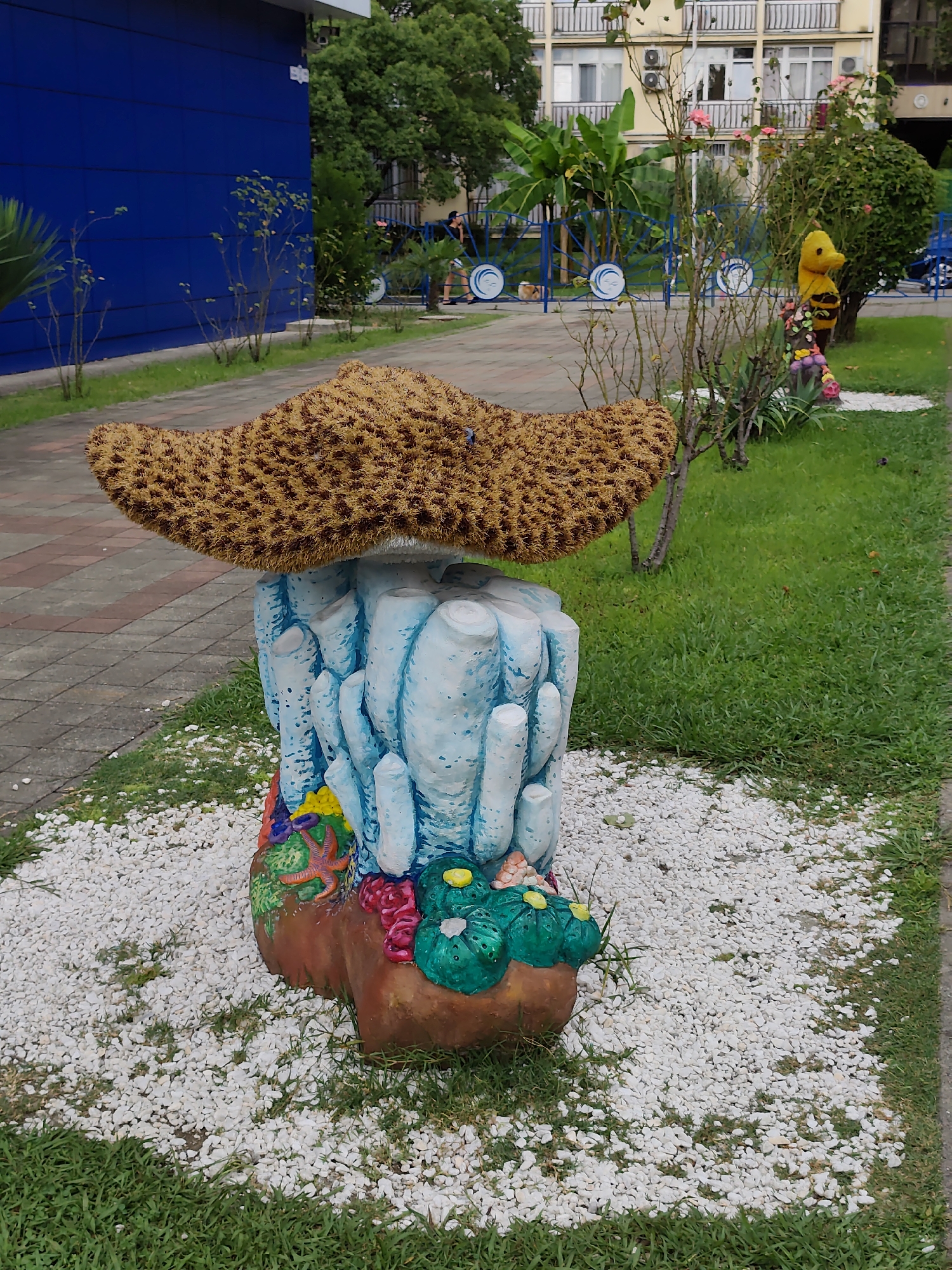 A garden sculpture resembling a starfish atop a coral base sits on a white pebble path in front of a blue building. In the background, a multi-story building with balconies and air conditioning units is visible, along with trees and a playground. A yellow bear figure is also present near some rose bushes. The scene appears to be in a public outdoor space, possibly a park or a hotel garden in Sochi, Russia.