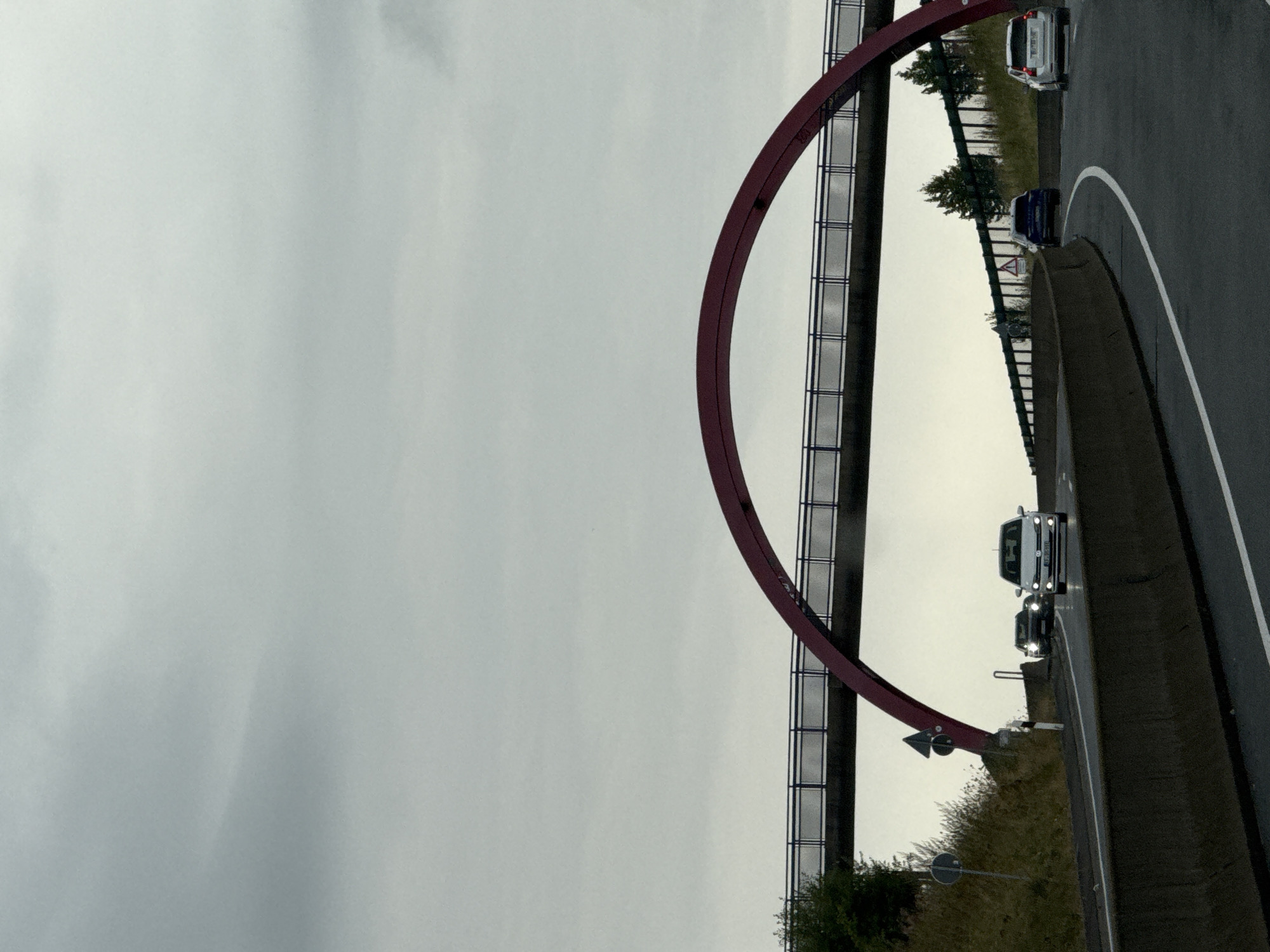 The image shows a highway under a distinctive red arched overpass in Bad Lauterberg im Harz, Germany. The sky is overcast and a light rain appears to be falling, indicated by the wet road surface and the headlights of approaching cars. Several cars are visible on the highway, traveling in the same direction. The overpass, designed as a pedestrian bridge, spans across the highway. The scene is captured from a moving vehicle, suggesting the photographer is a passenger. The surrounding environment is a mix of vegetation and highway infrastructure.
