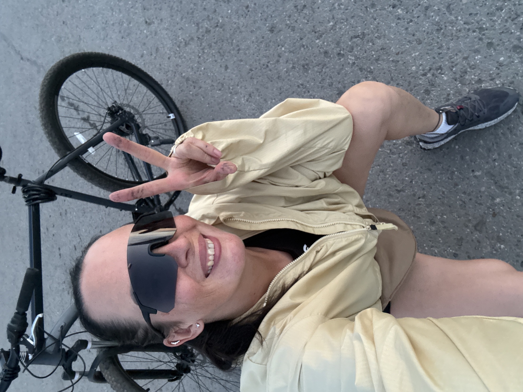A woman is crouching next to a bicycle, holding up two fingers in a peace sign. She is wearing sunglasses, a beige jacket, and tan shorts. The bicycle is black and has its front wheel turned. The background appears to be a paved road with scattered gravel. There is no identifiable visual information within the image to suggest a specific location in Chaikovsky, Russia.