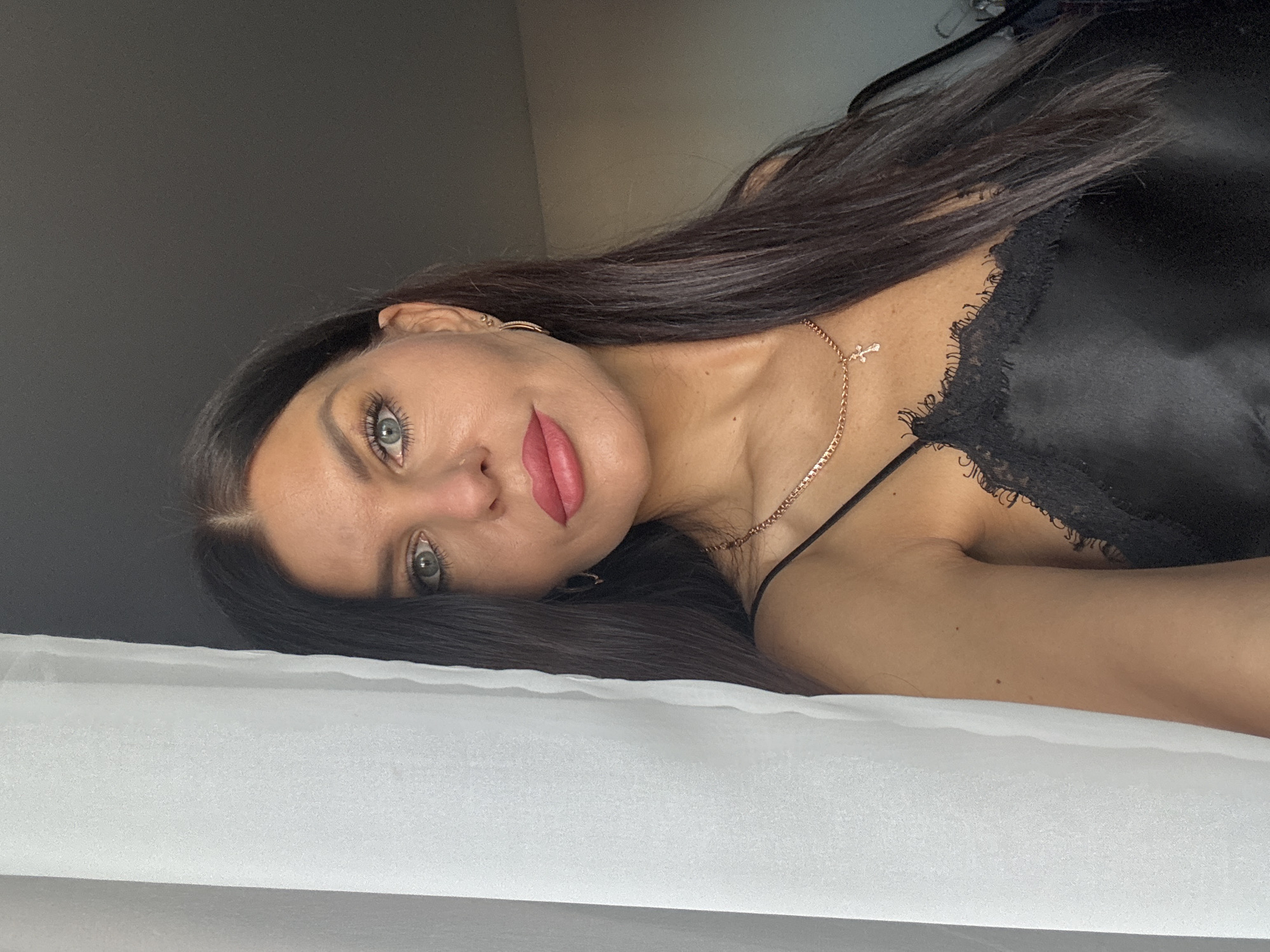 A portrait-style image captures a woman lying down, looking towards the camera. She has long, dark, straight hair flowing across the white surface she is resting on, and striking light-colored eyes (likely blue or grey). Her lips are adorned with pinkish-red lipstick, and her eyebrows are well-defined. She is wearing a black camisole or slip with a delicate lace trim visible along the neckline. A thin gold-colored chain necklace with a small cross pendant is around her neck, and small gold hoop earrings are visible in her ears.
The woman's head and upper body fill the left side of the frame, angled as if she is looking up from a reclined position. She has a calm and slightly smiling expression, with a direct gaze. Her head rests on a clean white surface, likely bedding, which occupies the bottom portion of the image. The background behind her is a plain, dark gray or black wall. The setting appears to be a private indoor space, possibly a bedroom. There is no visible text. The location is provided as Chaikovsky, Russia, though no specific visual cues within the image confirm this.