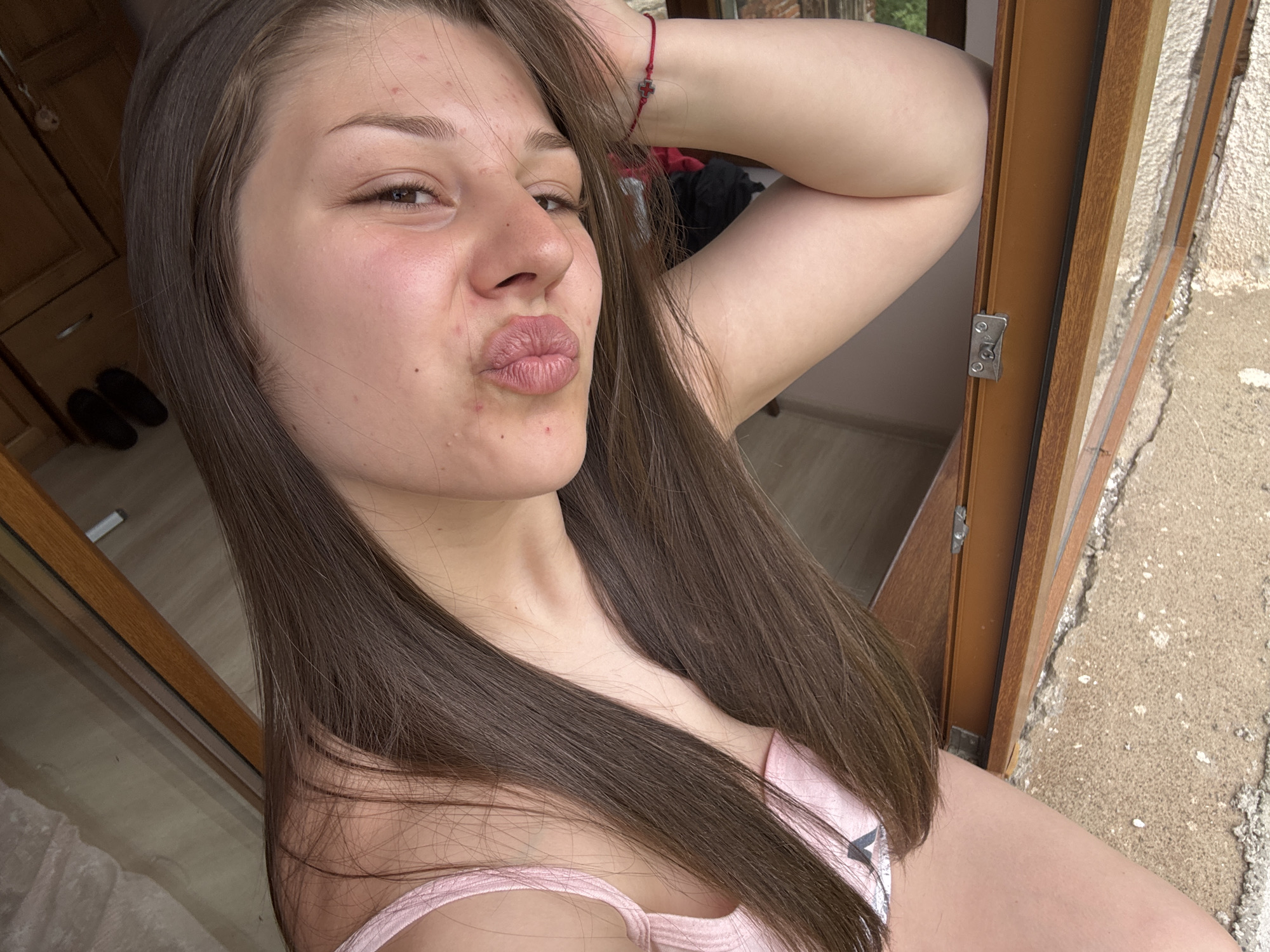 A young woman with long, brown hair is taking a selfie. She is leaning back, with her right arm raised and her hand on her head. Her lips are pursed, as if she is about to kiss the camera. She is wearing a light pink top. The background is out of focus, but it appears to be an indoor setting with wooden furniture and a window. The lighting suggests it is daytime.