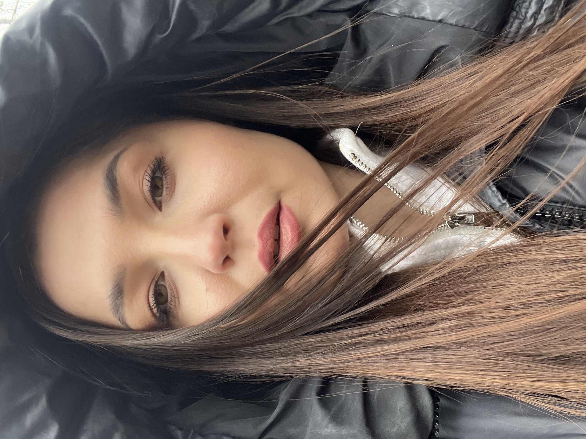 A woman with brown hair poses for a selfie. She wears a black hooded jacket over a white zip-up top. The location cannot be determined from the image.