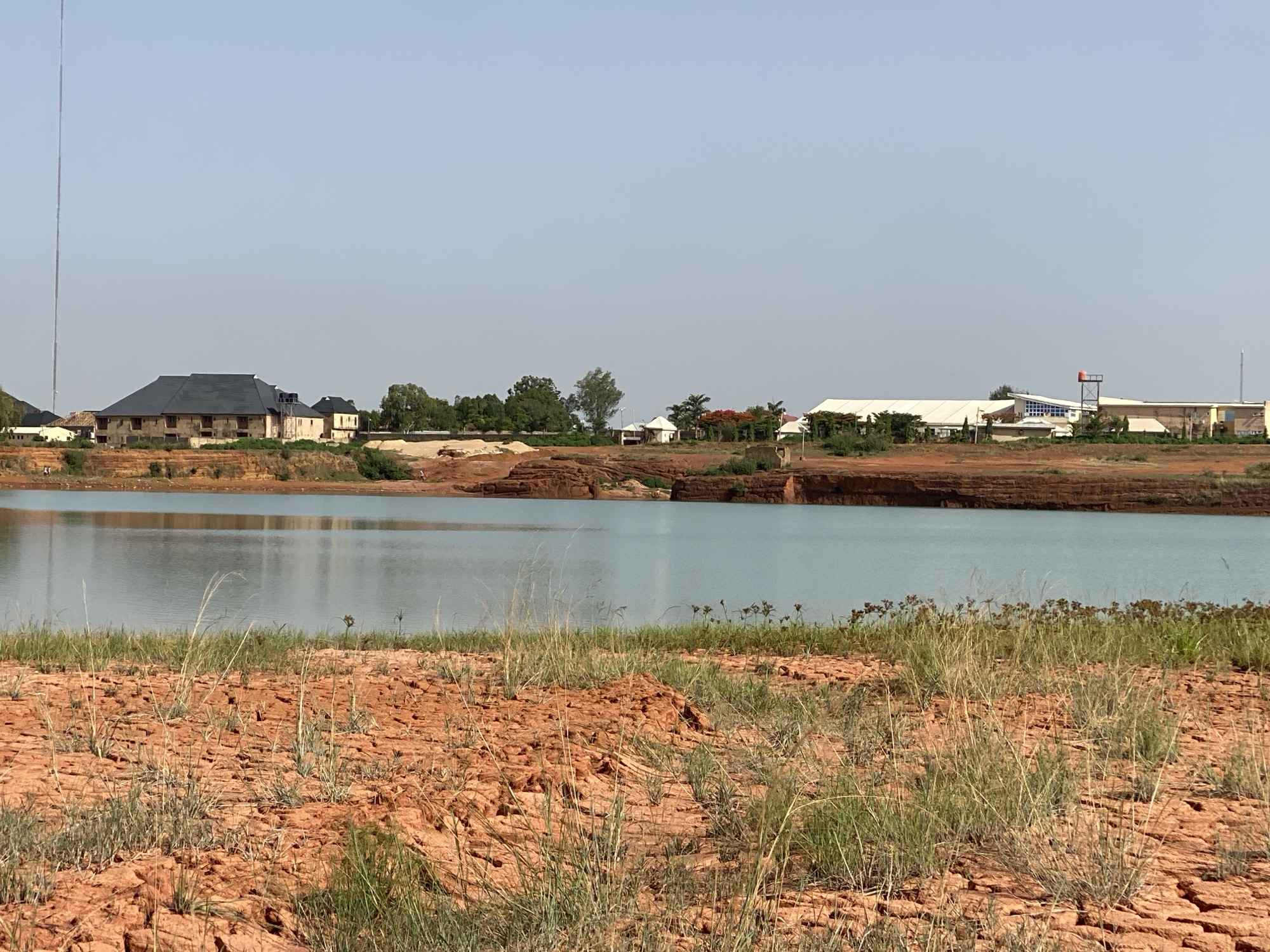 The image depicts a tranquil scene with a body of water in the foreground, possibly a lake or large pond, and structures of varying sizes and designs on the opposite bank. The foreground is characterized by dry, cracked, reddish-brown soil with sparse patches of green vegetation.
The water's surface is relatively calm, reflecting the sky and distant objects. Beyond the water, a range of buildings are visible, including what appears to be a large, multi-story building with a dark roof and light-colored walls, as well as several smaller buildings and structures. In the background, a tall tower or antenna rises into the sky.
The location appears to be in a rural or semi-urban setting, given the presence of both natural elements and man-made structures. It could potentially be the city of Unknown, Unknown. There are no people visible in the image. The sky is clear and pale blue, indicating fair weather conditions.