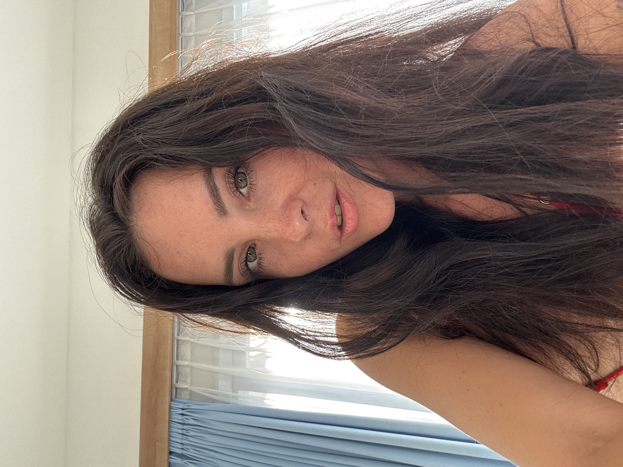 This is a close-up, low-angle, selfie-style shot of a woman with long, dark, wavy hair. She is looking towards the camera, with her face slightly tilted. Her eyes are green and her lips are slightly parted. She has a scattering of freckles across her nose and cheeks. Sunlight is streaming in from the right side of the frame, illuminating her hair and face. Behind her, a window with blinds is visible, and a blue curtain is draped across the bottom of the image. The overall impression is casual and intimate, possibly taken at home. The time of day appears to be daytime, with bright natural light. There is no visible text in the image. The context provided places the scene in Mengwi, Indonesia.