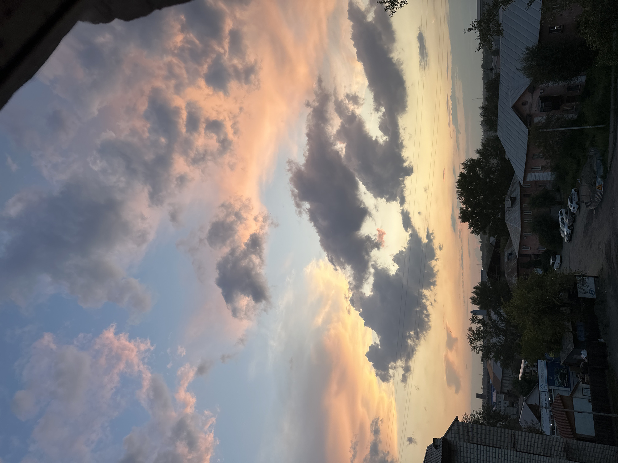 This is a photograph of a residential area in Biysk, Russia, taken during sunset. The sky is filled with dramatic clouds, tinged with shades of pink, orange, and gray. Below the sky, the rooftops of several buildings, including apartment blocks and smaller houses, are visible. Trees are scattered throughout the scene, and a few cars are parked on a road. The overall impression is one of a quiet evening in a moderately developed urban landscape.