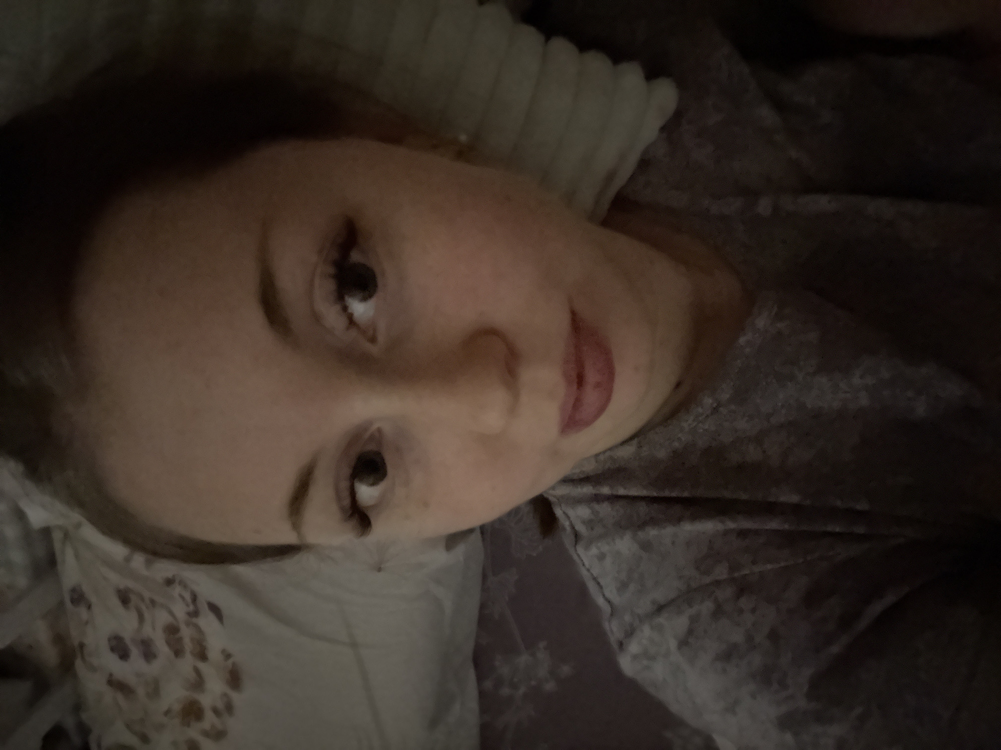 A selfie-style image depicts a person lying down, likely indoors. The person has fair skin, brown hair, and prominent eyelashes. The individual is positioned on what appears to be a pillow with a patterned case. Part of a grey, textured fabric, possibly clothing or a blanket, is visible. The location is unknown.