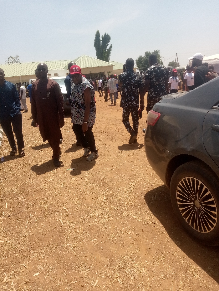 The image depicts an outdoor daytime gathering in Kagoma, Nigeria, on a flat expanse of reddish-brown dirt sparsely covered with dry straw-like debris. Bright sunlight creates distinct shadows across the ground. In the right foreground, a dark-colored sedan is partially visible, showing its rear passenger side and a multi-spoked wheel rim. In the mid-ground, from left to right, a man in a dark blue shirt is partially visible; a man in a brown tunic and dark trousers stands facing slightly left; and a man in a patterned short-sleeved shirt, dark trousers, and a red baseball cap with white lettering looks towards the camera. He wears light-colored sneakers. A small piece of light-colored litter lies on the ground near their feet. To their right, two individuals in dark camouflage uniforms walk away from the camera, moving towards a substantial crowd of people visible in the background. A light-colored building with a green roof and several trees are visible beyond the crowd. A light-colored vehicle is partially obscured behind the man in the brown tunic.