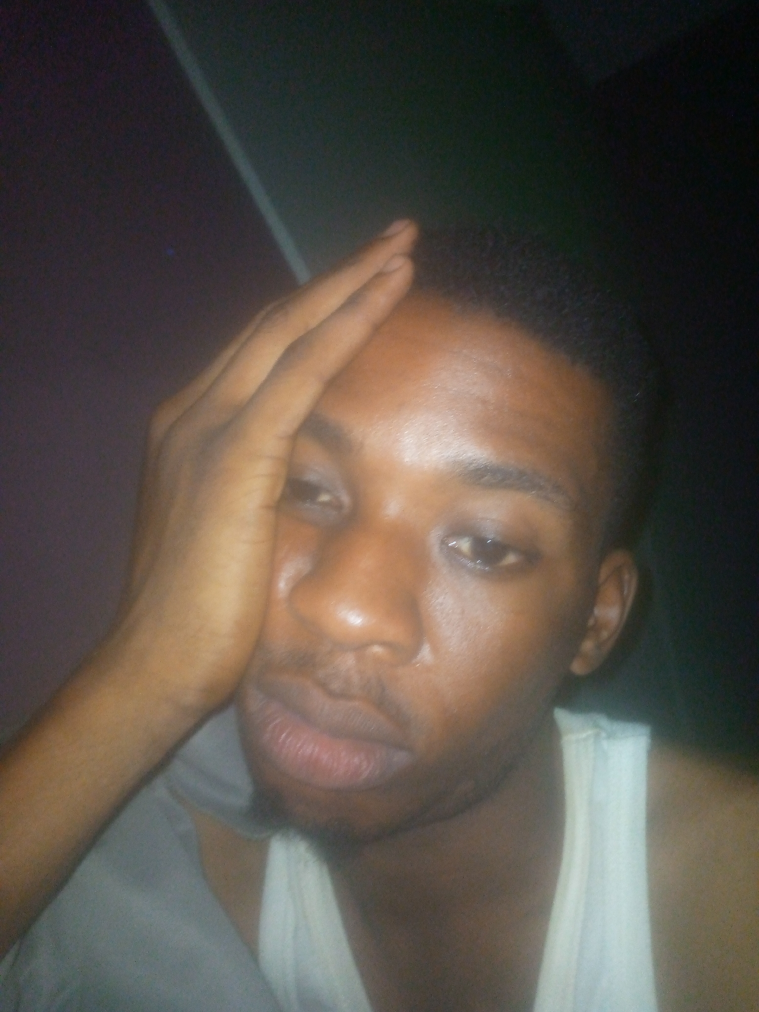 A man is pictured in an indoor setting with a dark, undefined background. He is wearing a white tank top and appears to have a gray shirt or cloth draped over his left shoulder. His right hand is positioned on his forehead, suggesting possible stress or discomfort. The location is unknown.
