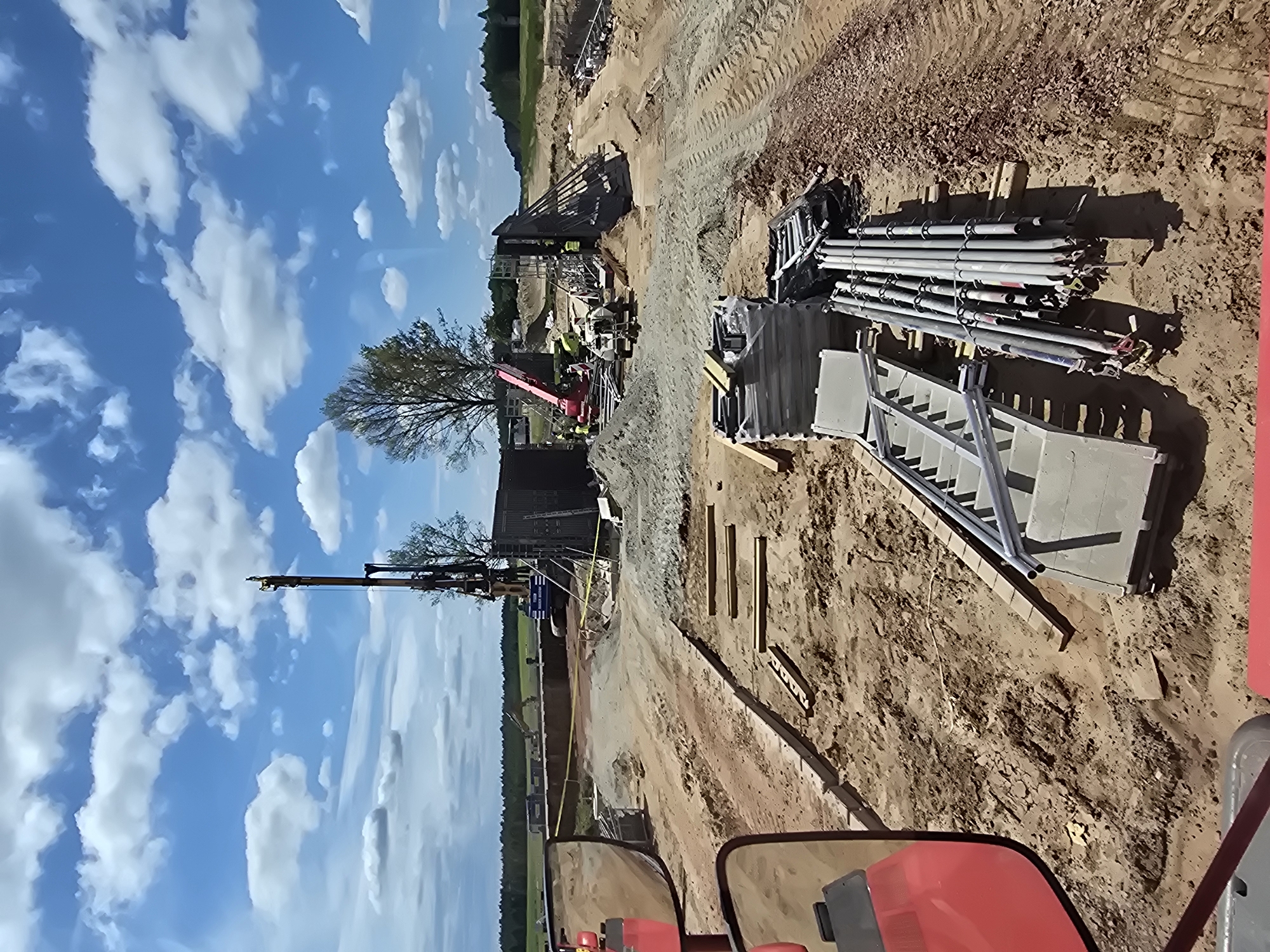The image depicts a construction site in Piecnik, Poland, under a bright, partly cloudy sky. A large drilling rig stands prominently in the background, suggesting deep foundation work or soil exploration. Adjacent to it, a red crane is visible, hinting at ongoing heavy lifting operations. The ground is uneven and consists of disturbed earth and piles of sand and gravel, typical of an active construction zone. Several stacks of metal scaffolding and construction materials, including pipes and structural elements, are organized in the foreground, indicating preparation for or progress in building. The scene appears to be a large-scale infrastructure project, possibly for a bridge or a significant building, given the scale of the equipment and materials. The overall impression is one of active construction and industrial activity in a semi-rural or suburban setting.