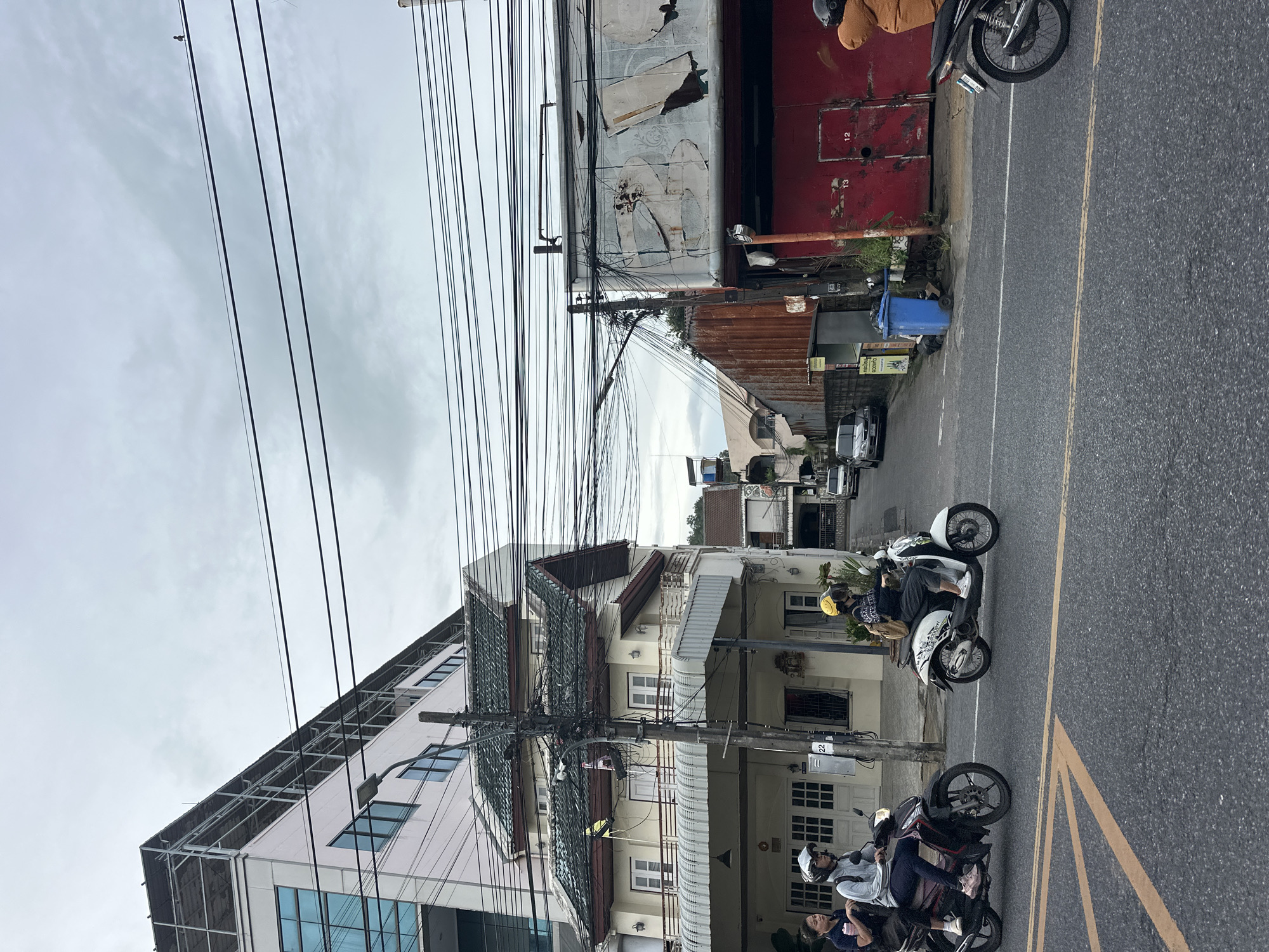 This is a vertical, eye-level shot of a street scene in what appears to be an urban area in Phuket City Municipality, Thailand. The image is dominated by a dense network of overhead electrical wires, creating a cluttered, almost abstract pattern against a cloudy, overcast sky. The buildings lining the street are a mix of styles and conditions, with some appearing modern and well-maintained, featuring glass windows and clean facades, while others show signs of age and disrepair, with rusted metal sheeting and peeling paint.
The street itself is paved asphalt with yellow lane markings. Two motorcycles are prominently featured in the foreground, each with riders wearing helmets. The activity appears to be typical street traffic, with people traveling on their motorcycles. One motorcycle, a white scooter, has two people on it; the driver wears a yellow helmet. The second motorcycle, a darker model, also has two people on it.
Notable details include a blue trash bin on the sidewalk and various signs with indiscernible text. The time of day appears to be daytime, given the lighting, but the overcast sky suggests it may be a cloudy or potentially rainy day, contributing to a somewhat muted color palette. The overall impression is one of a bustling, somewhat chaotic urban environment, typical of many Southeast Asian cities. The presence of numerous overhead wires is a distinctive characteristic of the scene.