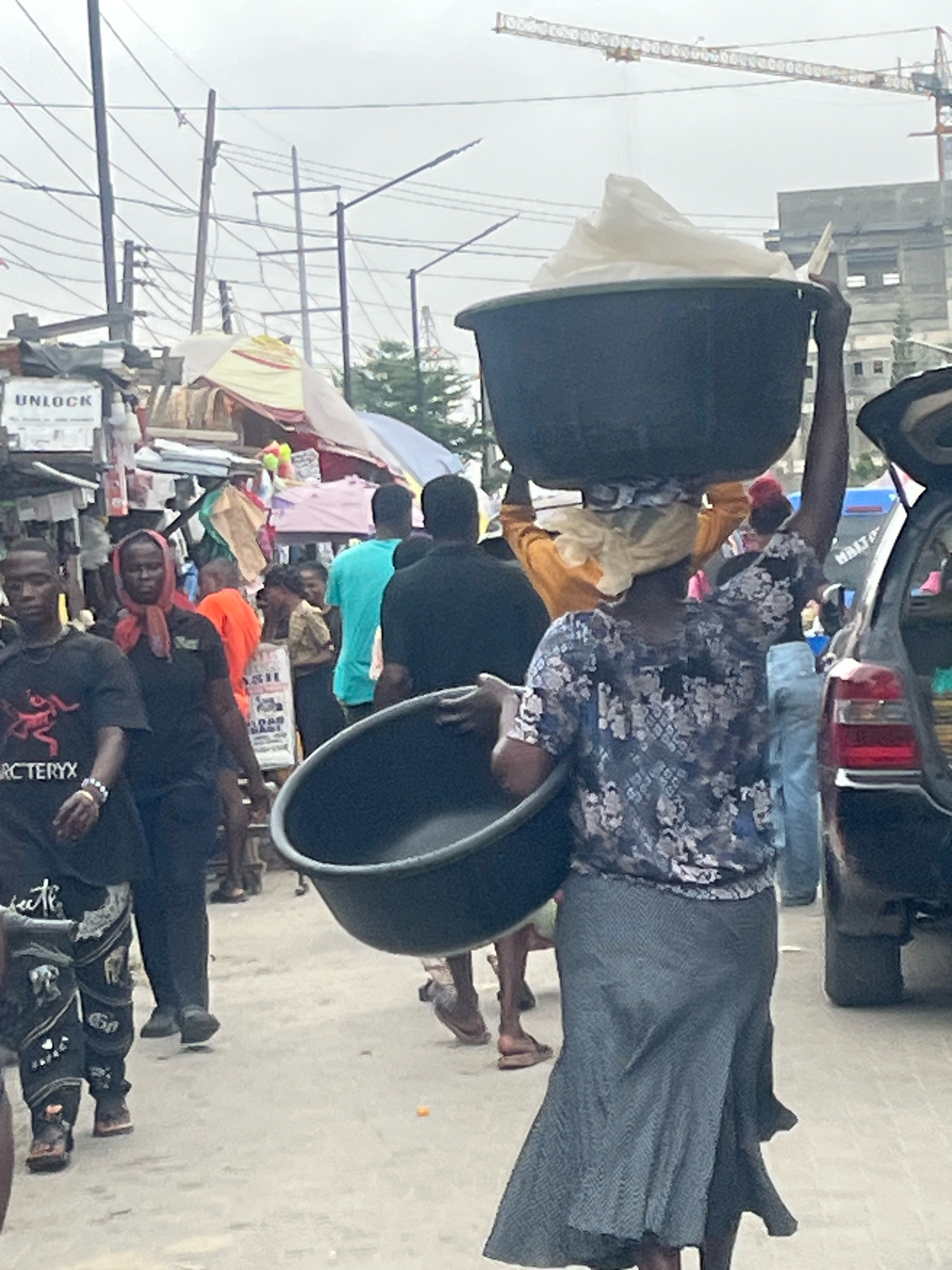 This image depicts a bustling street scene in Lekki Phase I, Nigeria, on an overcast day. In the foreground, a woman carries a large, dark basin filled with white material on her head. She wears a patterned blue and grey blouse and a grey skirt. Beside her, another person's arm is visible, also carrying a similar basin. To the left, a man in a black t-shirt with the "Arc'teryx" logo and patterned pants walks towards the camera. Behind him, another man in an orange shirt and a woman with a red headscarf are visible. In the background, various market stalls with colorful coverings, poles, and overhead electrical wires suggest a busy commercial area. A crane is visible in the far distance, indicating ongoing construction in the city. A dark-colored car is parked on the right side of the frame, with its trunk open. The ground is paved with concrete and appears to be a street or walkway.