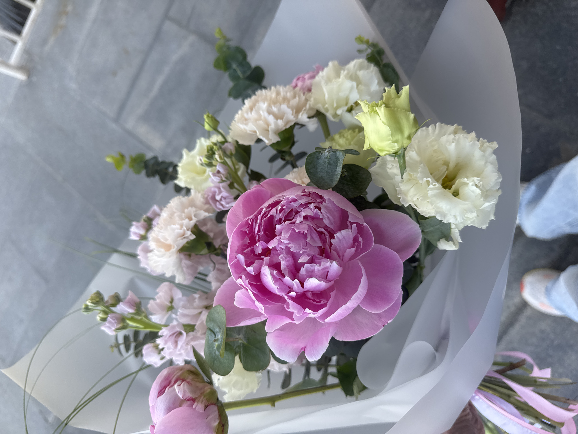 A bouquet of flowers, predominantly pink and white, is displayed. A large pink peony dominates the center of the frame. White carnations and other white flowers are interspersed throughout the bouquet. Green eucalyptus leaves are also present. The bouquet is wrapped in a translucent white material. The background is a grey tiled surface. A pair of sneakers are visible in the bottom right corner. There is no discernible evidence to confirm the location of Izhevsk, Russia.