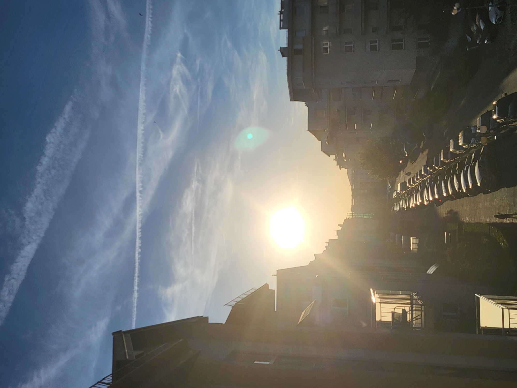 The image depicts a street scene in an urban area, likely at sunrise or sunset, as indicated by the bright sun obscured by buildings. Several multi-story residential buildings line the street, with parked cars on the right side. Vapor trails are visible in the blue sky to the left.