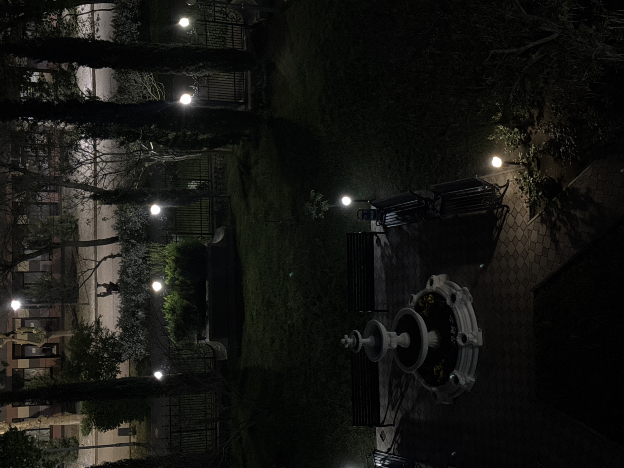 The image depicts a nighttime scene in a park in Yevpatoriya, Ukraine, possibly from an elevated vantage point. Key features include a fountain, benches, manicured lawns, trees and bushes, decorative fencing, and streetlights illuminating the scene. A person appears to be walking on a path in the distance. The buildings are visible in the background, on the other side of the park. The overall impression is one of an orderly, quiet, and well-maintained public space at night.