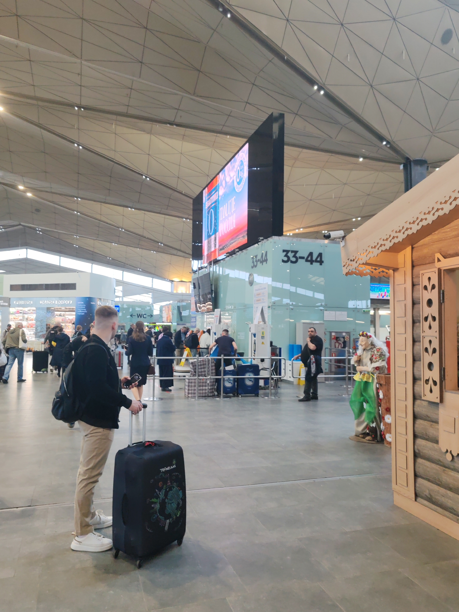 This image depicts the interior of a modern airport terminal, likely Pulkovo Airport in Saint Petersburg, Russia, characterized by its distinctive, geometric, light-colored ceiling. The scene is busy with travelers and airport activity.
In the foreground, a man with light brown hair, wearing a black jacket, khaki pants, and white sneakers, stands facing away from the camera. He is holding the handle of a black suitcase covered with a "TRAVELUUKS" protective cover, which features a globe and floral graphics. He appears to be engaged with his smartphone.
The background reveals a bustling airport environment. To the left, other passengers are visible near what seems to be a check-in or security area. A large, vertical digital display screen dominates the central area, showing text in Russian, including "ТОЛЬКО РОССИЯ" (Only Russia), "ТАНЦЕВАТЬ" (To Dance), and "0%". Below the screen, signs clearly indicate gate numbers "33-44" and a "WC" sign. A man with a beard, possibly airport personnel, stands in the mid-ground near the gate area.
To the right, a prominent wooden structure, resembling a traditional Russian log house (izba) with decorative carvings, stands next to a life-sized folk character figure. This figure is dressed in colorful traditional attire, including a floral wreath, and holds a staff, strongly evoking images of Russian folklore figures. The overall lighting is bright and even, typical of a well-lit indoor space, suggesting active hours of operation. The combination of modern airport design and traditional Russian cultural elements is a notable detail.
