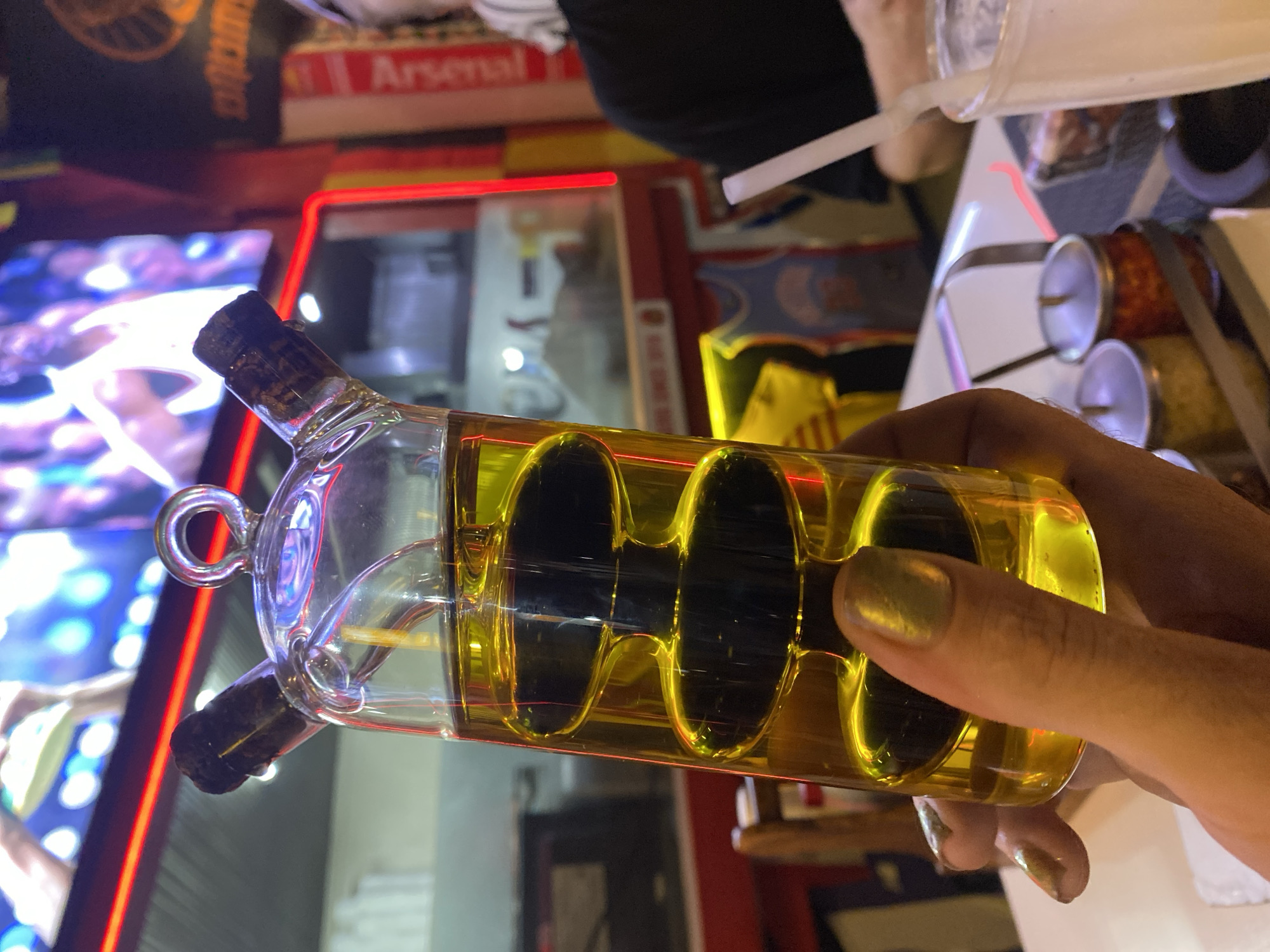 A person holds a glass oil and vinegar cruet in their right hand with gold-colored nail polish. The cruet is filled with a yellow-colored liquid and contains four inner glass segments that curve from one end of the bottle to the other. The bottle has two cork stoppers. The photo was taken at Moo 1, Thailand. In the background, there is a brightly lit Arsenal sign, a neon sign, and restaurant items.