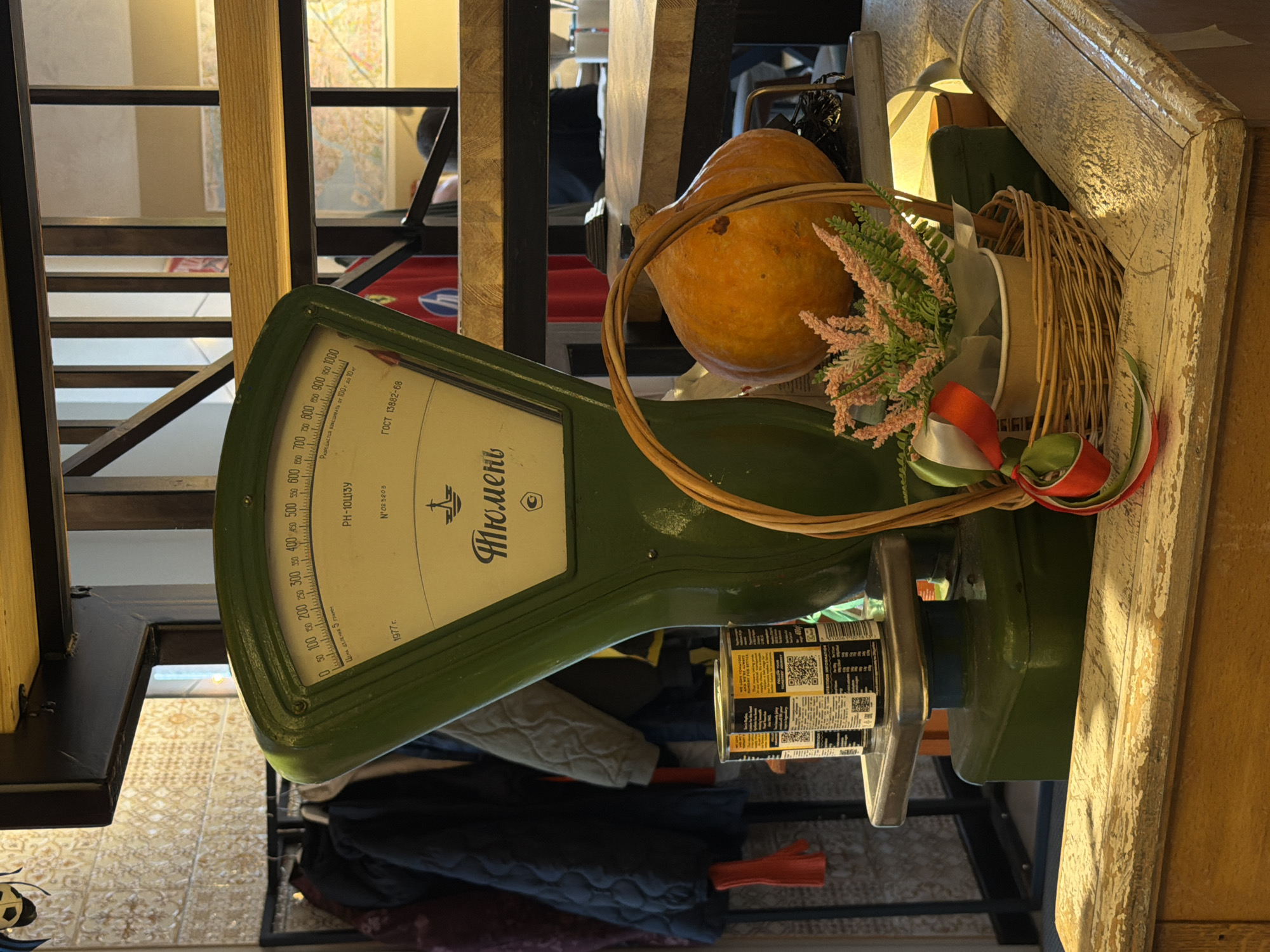 An antique green counter scale, rotated on its side, serves as a central display piece in what appears to be a rustic or vintage-themed interior in Izhevsk, Russia. The scale's dial, facing the viewer, shows measurements from 0 to 1000, with an inscription indicating "ЦЕНА ДЕЛЕНИЯ 5 ГРАММ" (division value 5 grams) and the manufacturing year "1977 г." (1977). Other Russian text includes "РН-10Ц13У," "N'023203," "Разрешается взвешивать от 100 г до 10 кг" (allowed to weigh from 100 g to 10 kg), "ГОСТ 13882-68," and the brand/city "Тюмень" (Tyumen).
Perched on the scale's platform is a large, vibrant orange pumpkin, next to a small wicker basket adorned with artificial pink and green floral arrangements and decorative ribbons (red, white, and green). A cylindrical item with black and yellow labels, featuring QR codes, is also visible on the scale.
In the background, a wooden and metal structure, possibly part of a staircase or shelving, creates depth. A person's head is partially visible behind these structures, along with a map on a distant wall. The foreground reveals a patterned tiled floor and what appears to be dark-colored coats or clothing hanging beneath the display. A rustic, distressed wooden beam frames the right side of the image, contributing to the cozy and traditional ambiance of the scene. No specific activity is depicted; it's a still life arrangement, likely serving as decoration.
