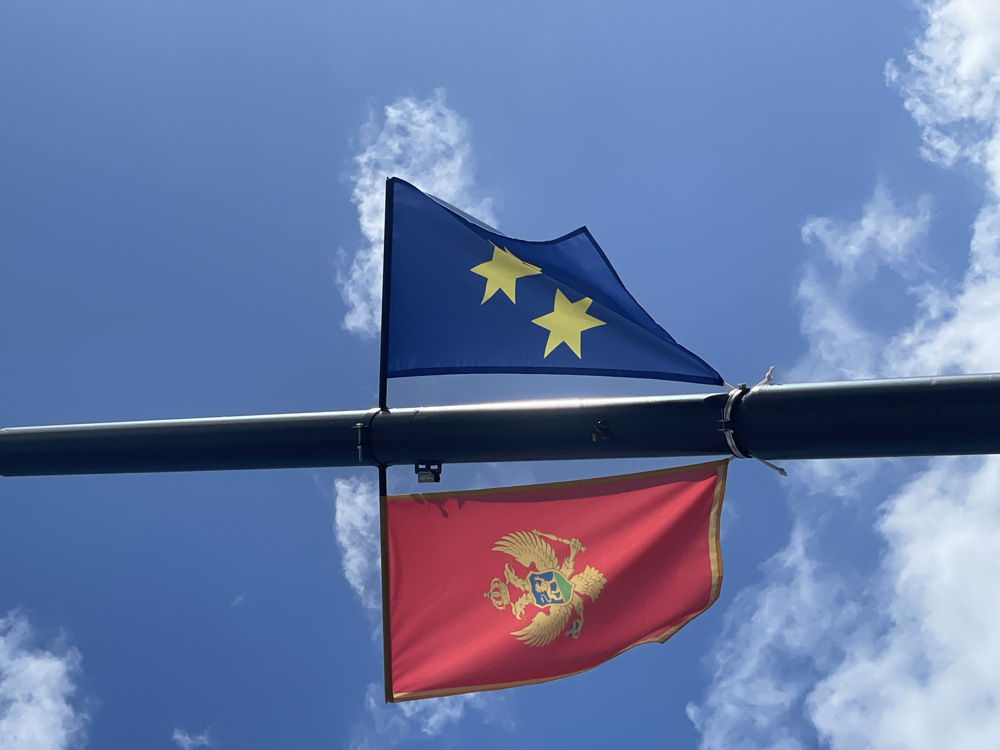 Two flags are affixed to a dark-colored horizontal pole against a backdrop of a blue sky with scattered white clouds. The upper flag is blue with two yellow stars. The lower flag is red with a gold emblem consisting of a double-headed eagle. Based on the flag designs, the upper flag is the European flag with two stars and the lower is the flag of Montenegro.
