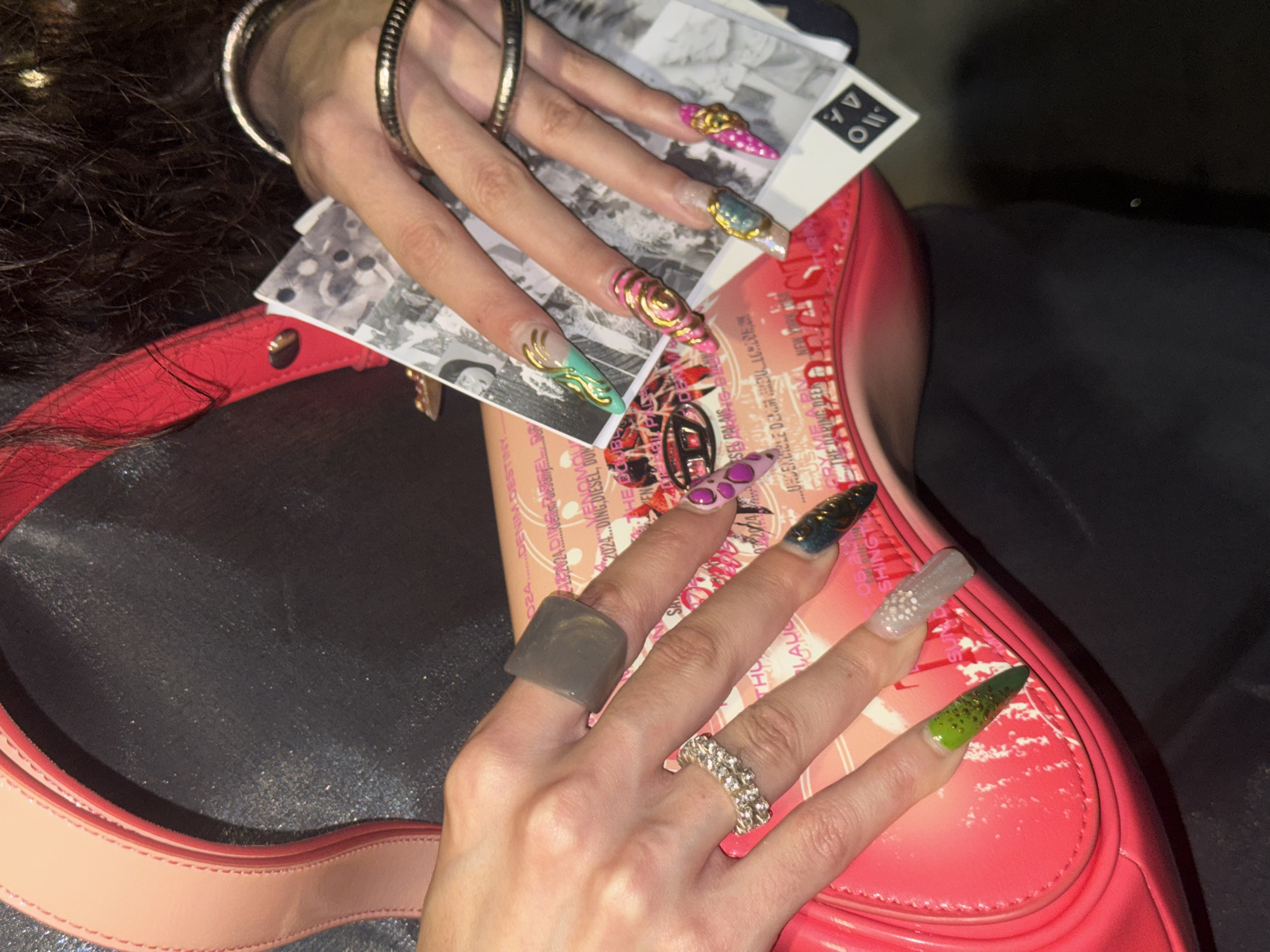 The image shows a close-up of a person's hands and a bright pink handbag. The person has long, elaborately decorated fingernails with various colors and designs, including glitter, metallic accents, and 3D embellishments. They are wearing several rings on their fingers. The handbag appears to be made of patent leather and has a printed pattern on it. The background is dark and out of focus, suggesting an indoor setting or low light conditions. There is no indication of the specific location within the city of Sofia, Bulgaria.