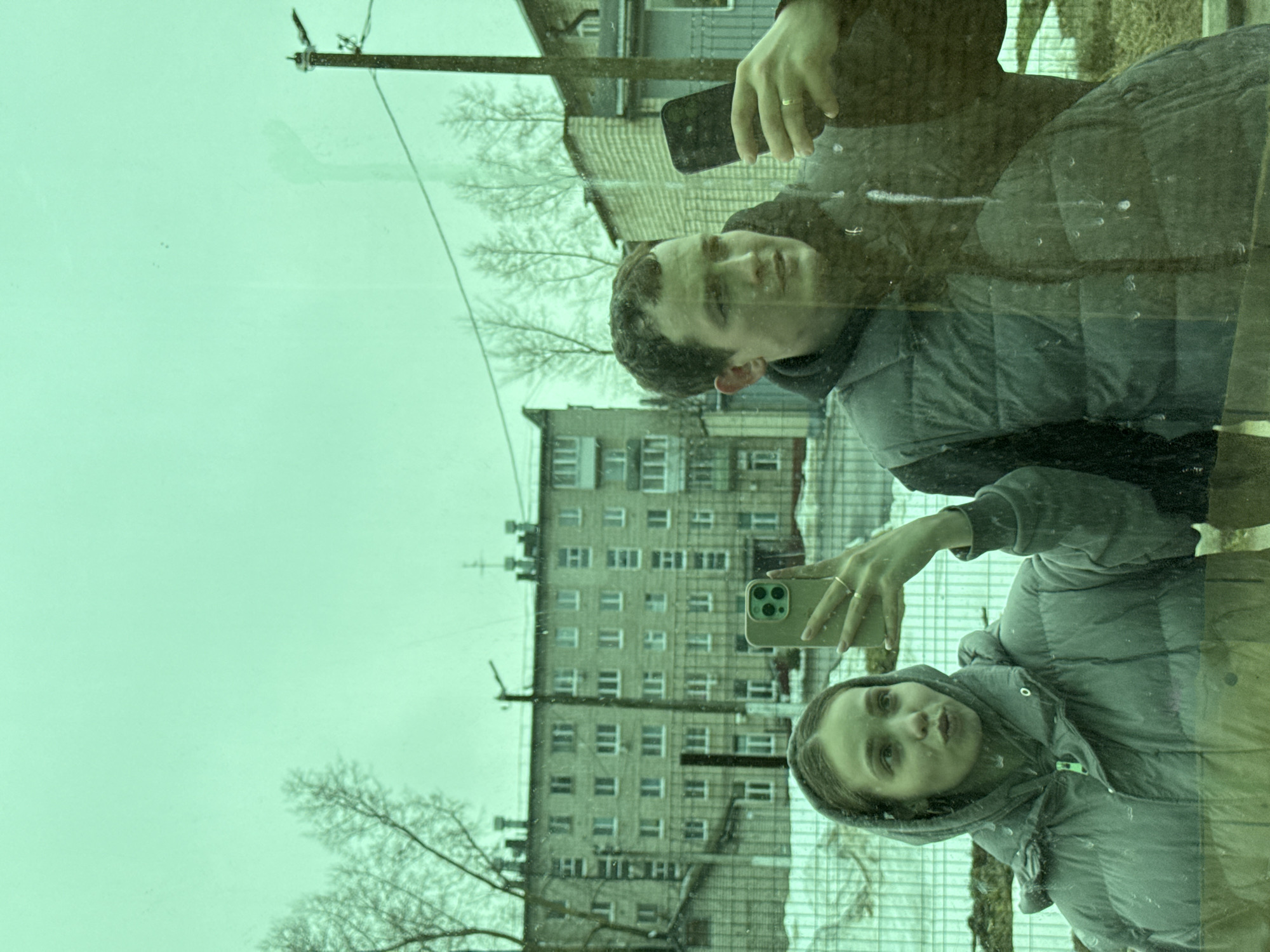 The image shows a selfie reflected in a window, with a building visible in the background. The building, likely located in Chaikovsky, Russia, has multiple floors with numerous windows. In the reflection, a young man is holding a black phone, and a young woman is holding a green phone. They appear to be taking a picture of themselves with the building in the background. The man wears a dark hoodie and grey puffer jacket, and the woman wears a grey hooded puffer jacket. A power pole and bare trees are visible in the background. The glass has a green tint and smudges.