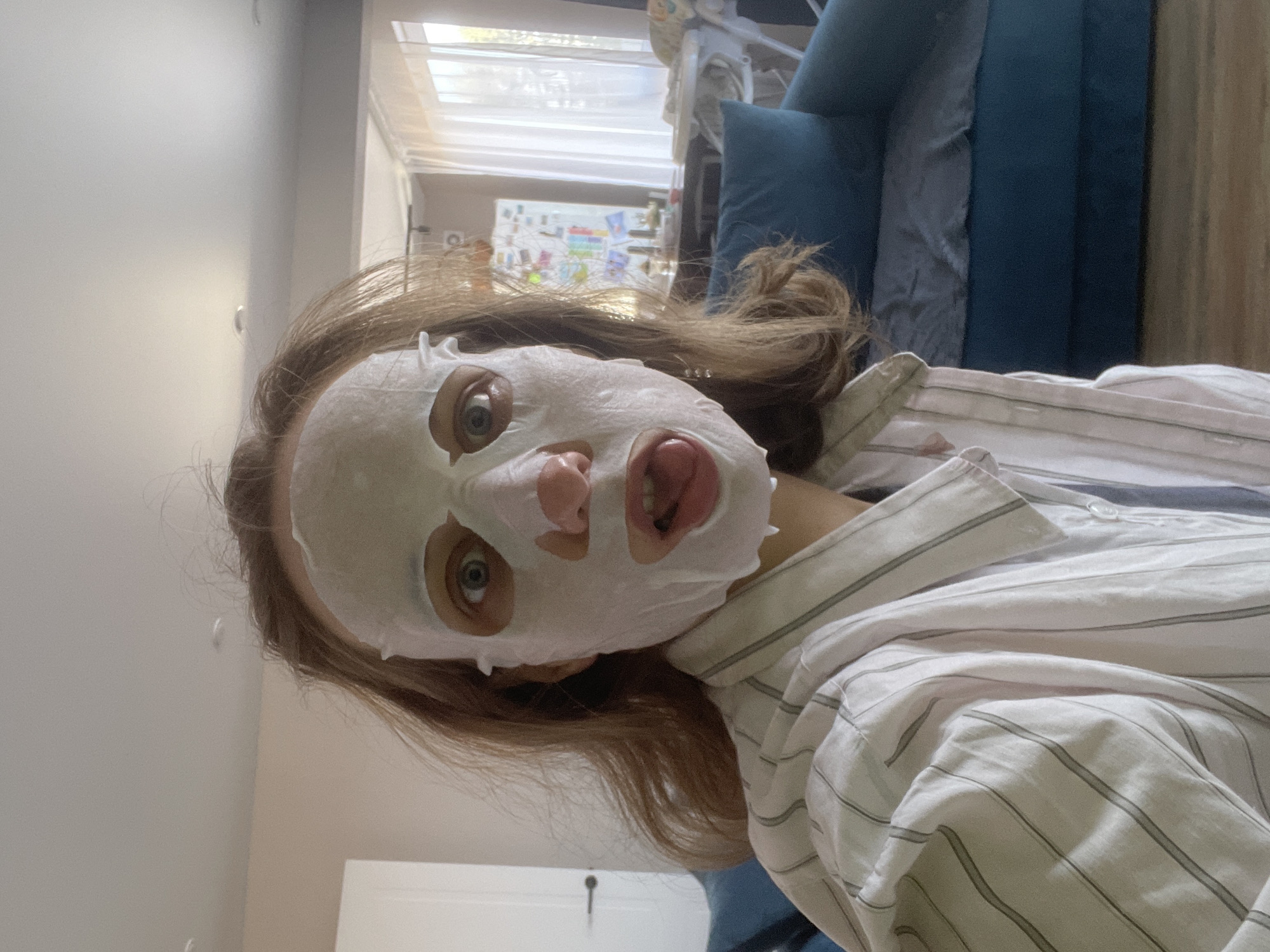 A woman wearing a white sheet mask is pictured in a vertical, close-up shot. Her eyes are visible through the mask's eye holes, and her mouth is slightly open. She wears a light-colored, striped button-down shirt. Her brown hair is loose and slightly tousled. The background includes a blurred view of what appears to be a room interior, including a blue sofa and a window with sheer curtains. In the background on the window are what appear to be colored papers and pictures that look to be from children. A white door is partially visible on the left side of the frame. The image was likely taken indoors in Sepych, Russia.