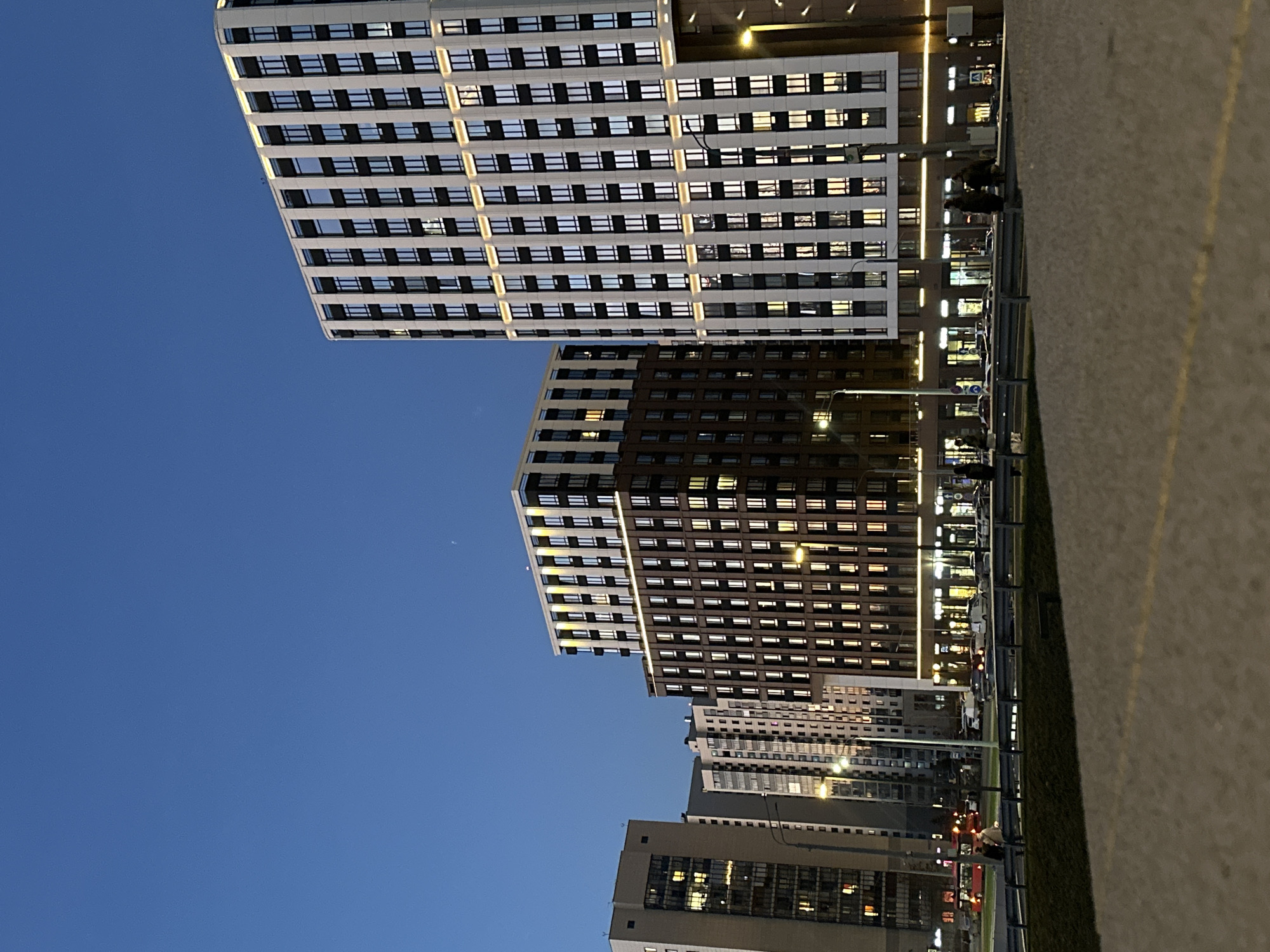 The image captures an urban evening scene, likely at twilight or early night, featuring multiple modern high-rise residential or commercial buildings under a deep blue sky. The photo is taken from a slightly low angle, looking up at the structures.
In the foreground and to the left, a tall building with a facade of white horizontal bands and dark rectangular windows dominates the view. Many of its windows are dark, while others glow with warm yellow light. Adjacent to and slightly behind it, a similar-style but darker, brown-colored building is prominent, also showing numerous illuminated windows. Further in the background and to the right, several other multi-story buildings can be seen, some lighter in color, contributing to a dense urban skyline, all with various windows lit up.
The ground level features a wide, paved area, likely a sidewalk or esplanade, with a faint yellow line running along its edge on the right. This paved area is separated by a low barrier or fence from a strip of greenery (possibly grass or low bushes). Beyond this, a street or parking area is visible with several vehicles, some parked and others possibly moving, indicated by their headlights or taillights. A few indistinct figures of people can be seen standing or walking near the barrier. Streetlights emit bright yellow light, illuminating parts of the scene.
The sky is a clear, dark blue, indicating the evening hour. A faint, small crescent moon is visible in the upper left portion of the sky. No discernible text is clearly visible on any signs or buildings. The overall atmosphere is one of a bustling city settling into the night, with the illuminated windows and streetlights providing a sense of activity and warmth against the cool evening sky.
