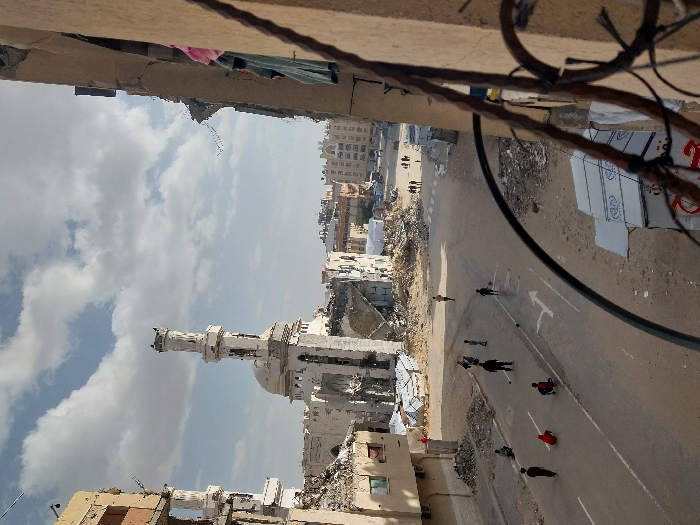 The image captures an elevated view of a street in Khan Yunis, Palestinian Territory, under a partly cloudy sky. In the mid-ground, a large mosque with a prominent dome and a significantly damaged minaret stands amidst extensive rubble and partially collapsed buildings. The street, an asphalt road with faded white arrow markings, is flanked by debris piles from destroyed structures.
Approximately 10-12 individuals are visible on the street. In the foreground-mid-ground, four individuals, including children and young adults, are playing with a dark soccer ball; two are standing and two are crouched. Other individuals are walking or standing further down the street, and a small group of five figures is visible crossing a distant crosswalk.
On the immediate right foreground, a damaged building wall partially obstructs the view, with articles of clothing (pink, light blue) hanging from an opening. A twisted metal railing extends across the lower right frame. To the left, a multi-story building shows signs of structural damage. Sheet metal panels with partial Arabic text are visible on the ground level to the right of the street. The surrounding urban environment is characterized by widespread structural damage and scattered debris.