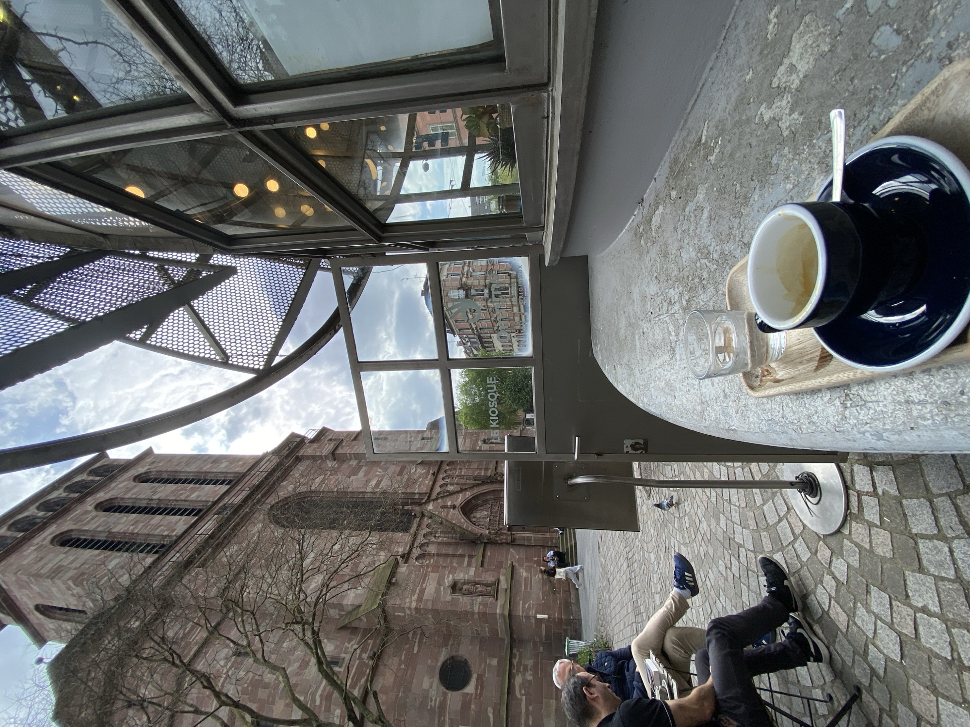 The image, taken from a low-angle perspective, depicts an outdoor cafe setting in Strasbourg, France. In the foreground, a cup of coffee sits on a table, with a small glass beside it. The table is made of a light grey, weathered material. In the background, the facade of a large, red brick building, presumably a church or cathedral, is visible. The building's architecture includes arched windows and detailed stone carvings. A bare tree stands in front of the building.
Several people are seated at the cafe, partially visible from the waist down. One person is wearing beige pants and blue sneakers, while another is wearing black pants and black sneakers. Other individuals are further in the background, near the entrance to the building.
Above the table, a metal structure with a glass roof is visible. The glass reflects the surrounding buildings and sky. A sign with the word "KIOSQUE" is visible in one of the glass panes. The ground is paved with cobblestones. The sky is cloudy.