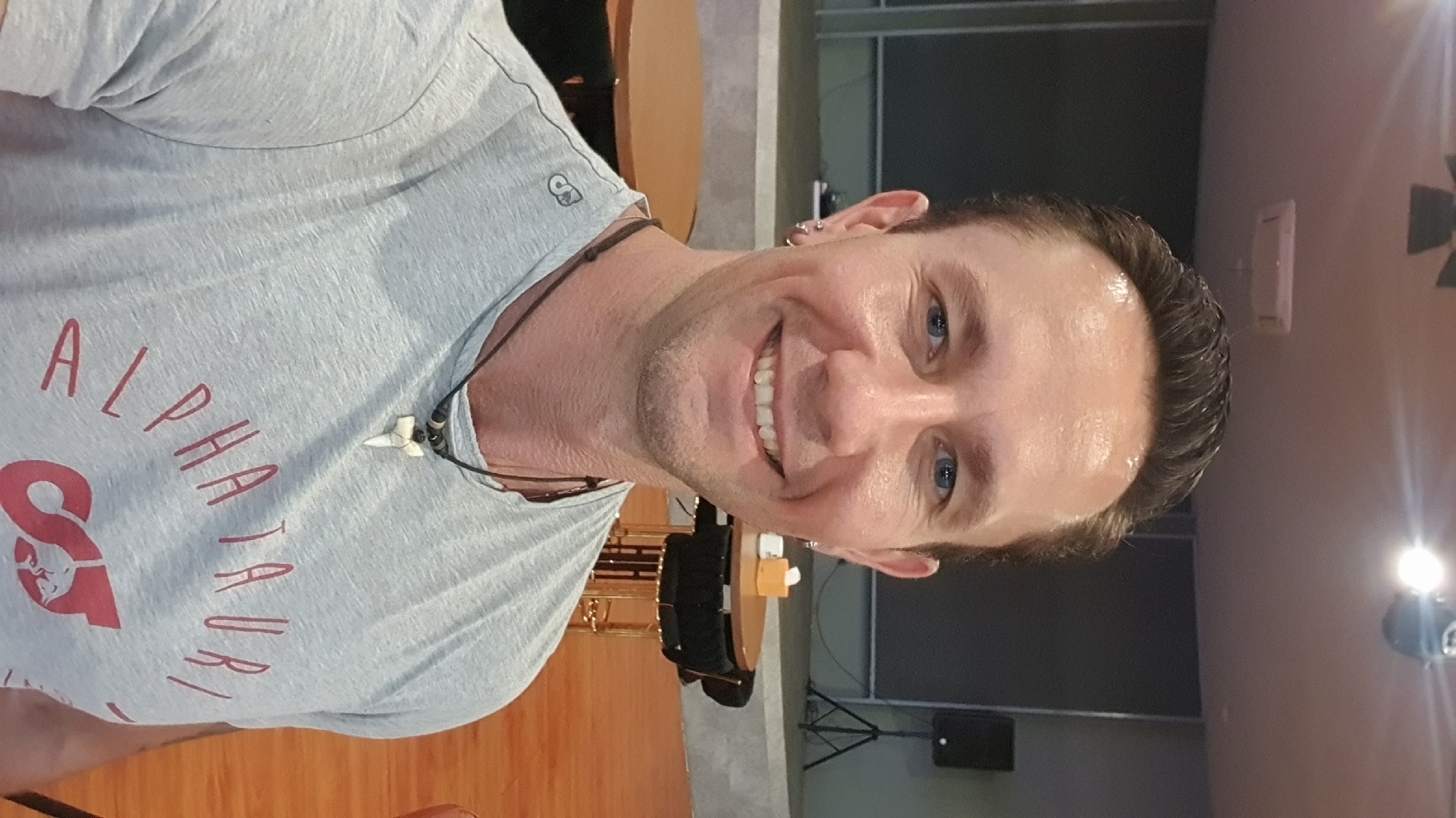 The media file shows a close-up, portrait-style, selfie-taken photograph of a man. He is wearing a light grey t-shirt with red lettering that reads "ALPHA TAURI". The man has short, dark hair, blue eyes, and is smiling, showing his teeth. He is wearing a necklace with a shark tooth pendant and some beads. The background appears to be indoors, with wooden floors and furniture visible. There is a speaker and some lighting equipment in the background, suggesting the setting could be a performance space or a meeting room. The lighting suggests it is likely daytime, or at least a well-lit indoor space. The overall mood of the photograph is casual and friendly, with the man appearing happy and relaxed. The image is oriented vertically, as if the person was holding the camera up to take a selfie. There is a small logo on the man's shirt, just above his left shoulder, which is a stylized "S" inside a circle.