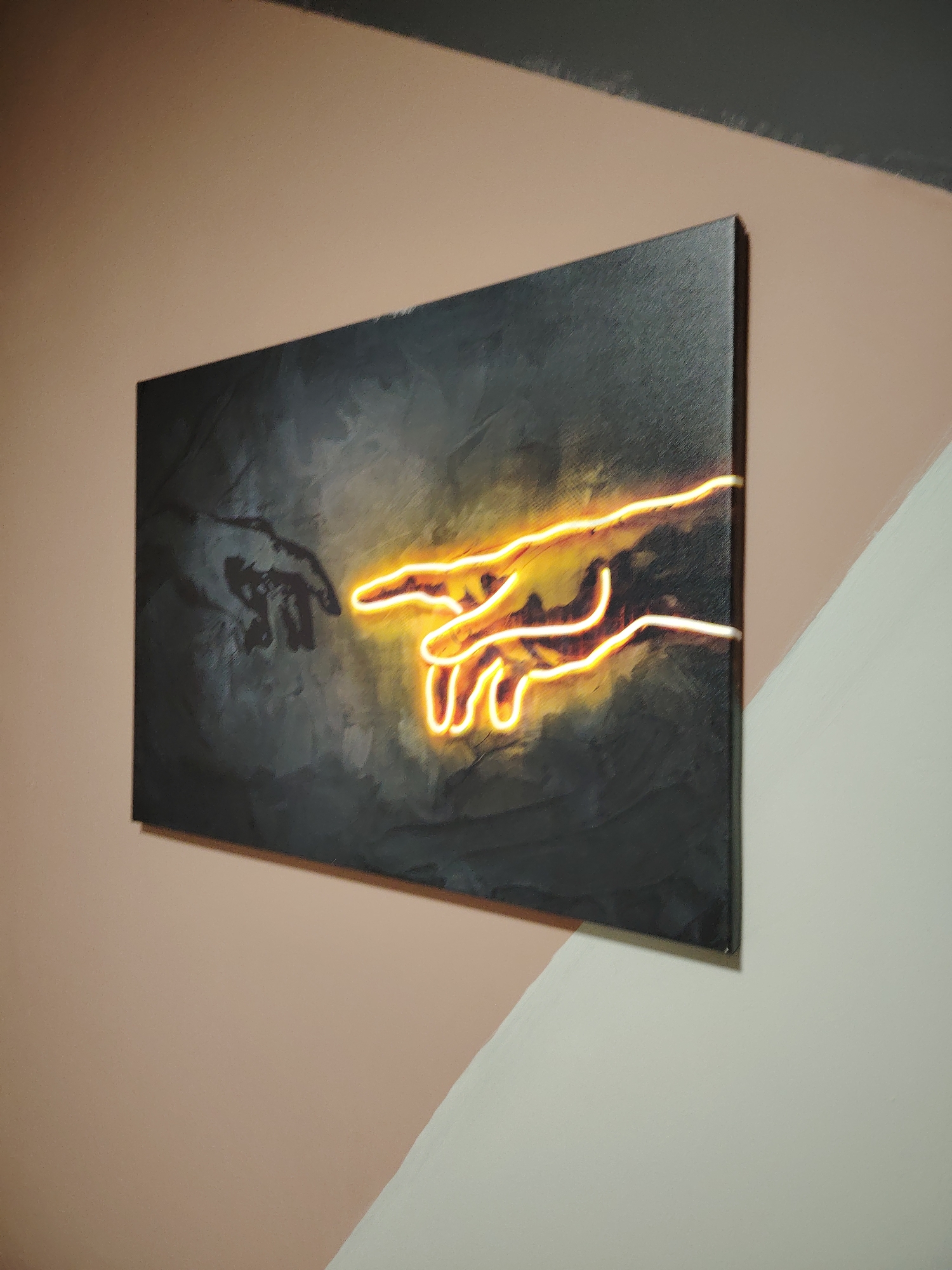 A painting depicting a hand silhouette reaching towards another hand that is illuminated in yellow and orange tones hangs on a wall that is painted in multiple colors. The top portion of the wall is dark gray, the middle is light pink, and the bottom is white. The location is Марковский, Russia.