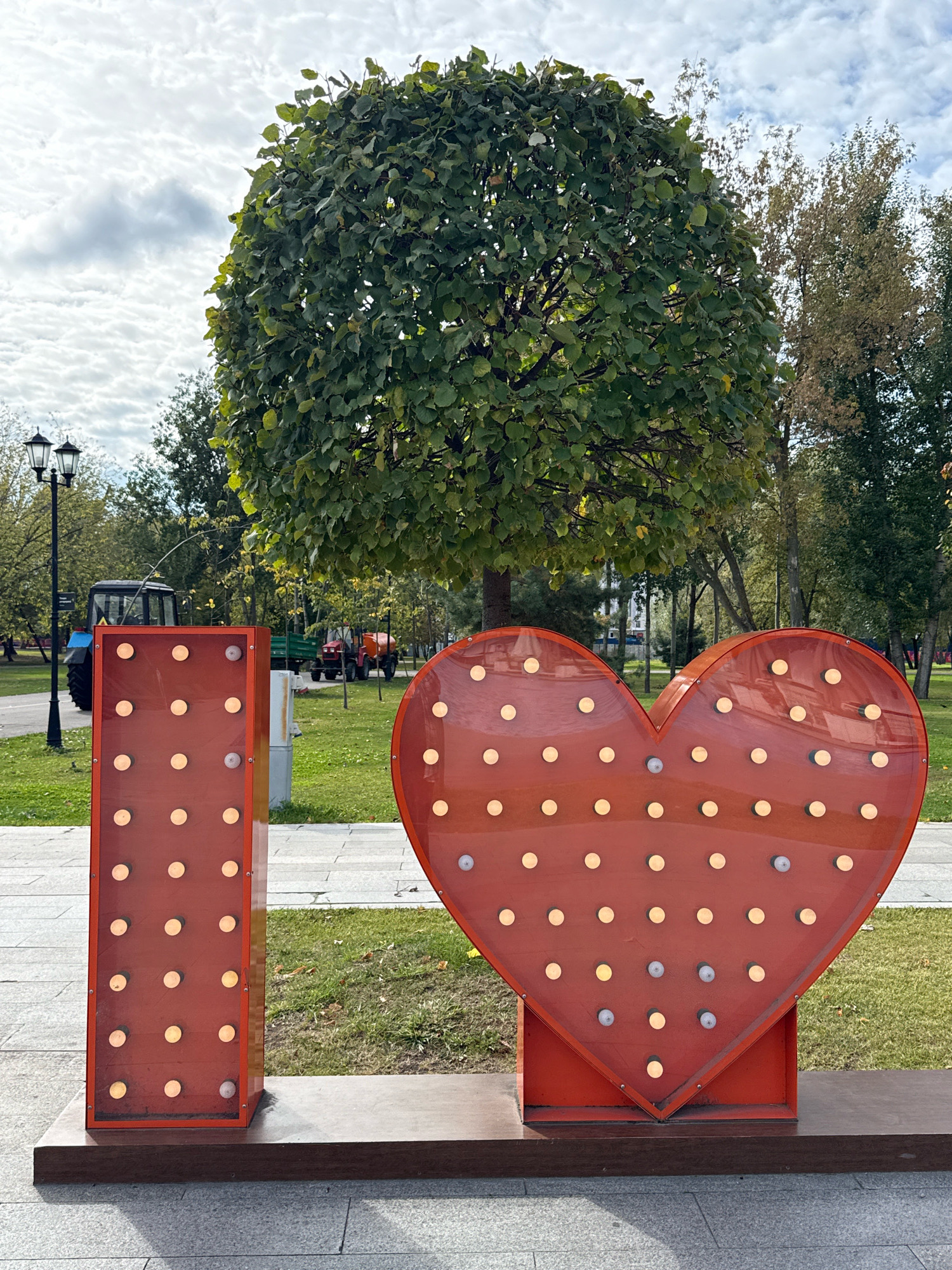 The image depicts an outdoor scene in a park, likely during the daytime. In the foreground, there are two large, illuminated decorative elements: a tall, rectangular structure with lights arranged in a grid, and a large, heart-shaped structure with lights similarly arranged. These elements appear to spell out "I ❤️". The structures are red and stand on a wooden base. Behind them, a large, spherical tree with lush green foliage is prominent. In the mid-ground, a paved pathway is visible, and beyond that, a blue tractor and a red tractor with a trailer are parked on a grassy area. Several other trees with green and some autumnal foliage are scattered throughout the background, along with lampposts. The sky is overcast with white clouds. The overall impression is of a public park setting, possibly decorated for an event or as a permanent installation. There are no people visible in the image.
