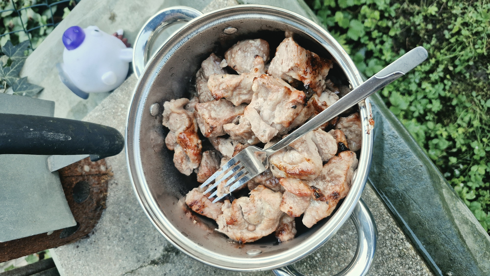 A metallic pot filled with grilled meat chunks and a fork is positioned on a concrete surface outdoors. In the background, a rusted charcoal starter, a dark handle, a small white toy pig with a purple top, and green foliage are visible. The scene appears to be in a backyard or garden setting, possibly on a patio. There are no people or discernible city landmarks.
