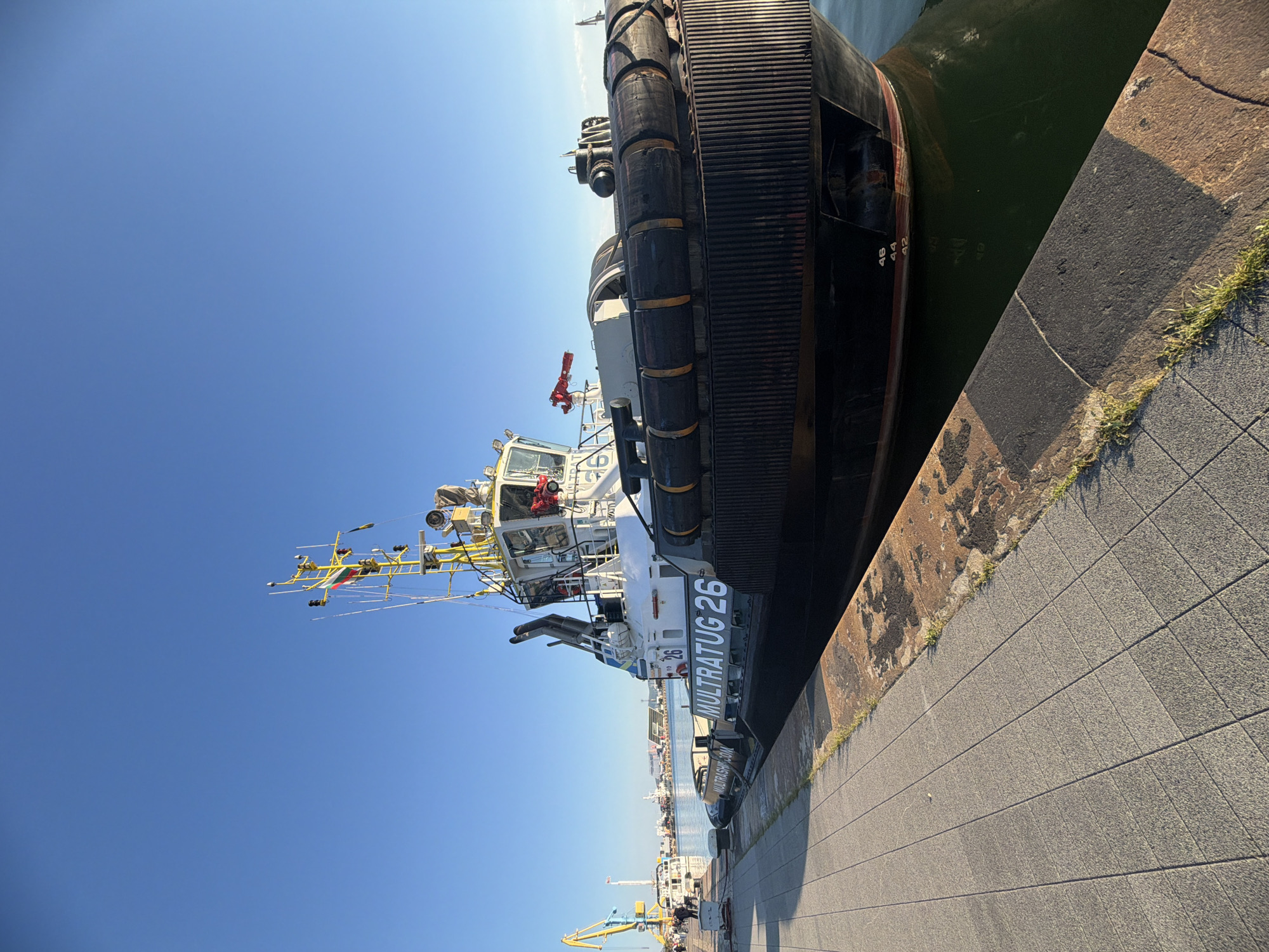 A daytime shot depicts a Multratug 26 tugboat docked at a pier in an unidentified city. The tugboat, painted with white and black sections, sits alongside a paved walkway. Its exterior is equipped with rubber fenders. The water separates the boat from the pier. In the background, harbor elements such as cranes and structures are visible. The sky is clear blue.