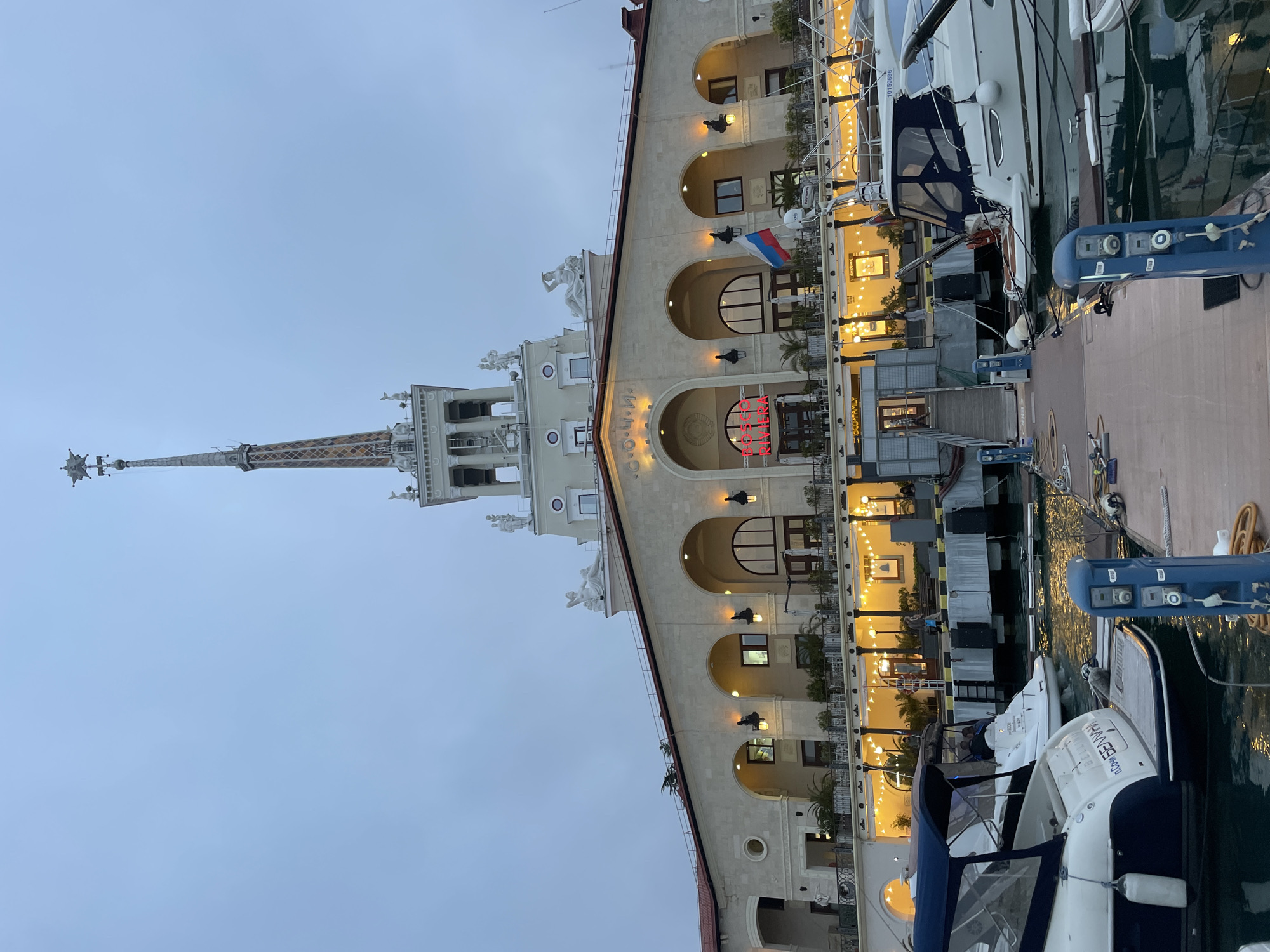 The image depicts the Sochi Marine Terminal in Sochi, Russia. It is an evening shot capturing the terminal building with its distinctive spire and classical architecture, adjacent to a marina. The building is illuminated, showcasing arched windows and ornate details. A Russian flag is visible on the facade. Several yachts are docked along the pier, positioned against the building. The sky is overcast.