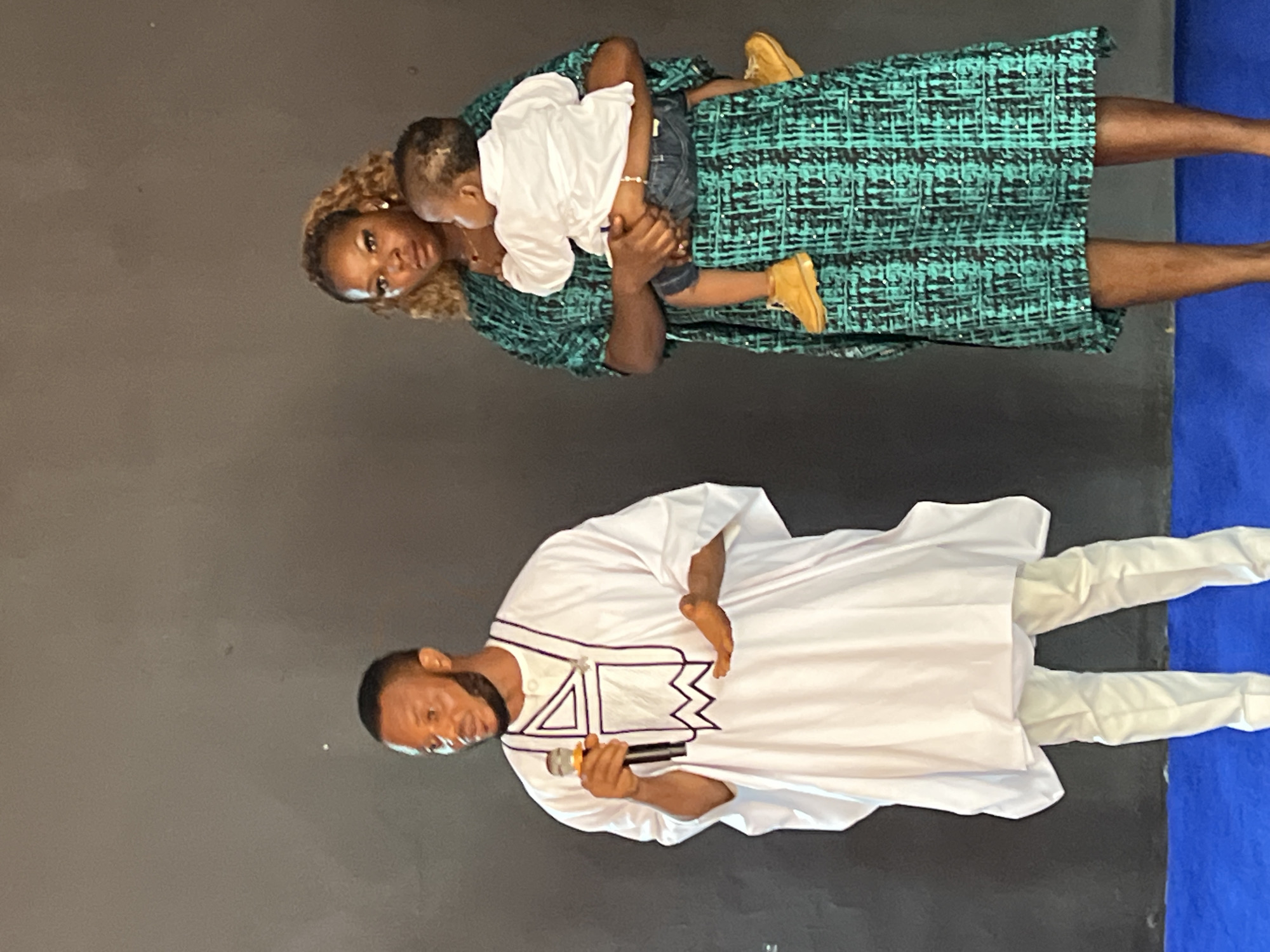 In Nnewi, Nigeria, three individuals stand against a dark backdrop, likely an indoor setting with a plain wall. On the left, a man dressed in a white kaftan with black trim holds a microphone and gestures with his right hand, suggesting he is speaking or performing. He has a beard and short hair. To his right, a woman in a teal and black patterned dress holds a young child. The child, wearing a white shirt and blue shorts, is facing away from the camera. The woman has blonde, curly hair and is looking towards the child. The floor in front of them is a vibrant blue. The lighting is moderate, casting some shadows.