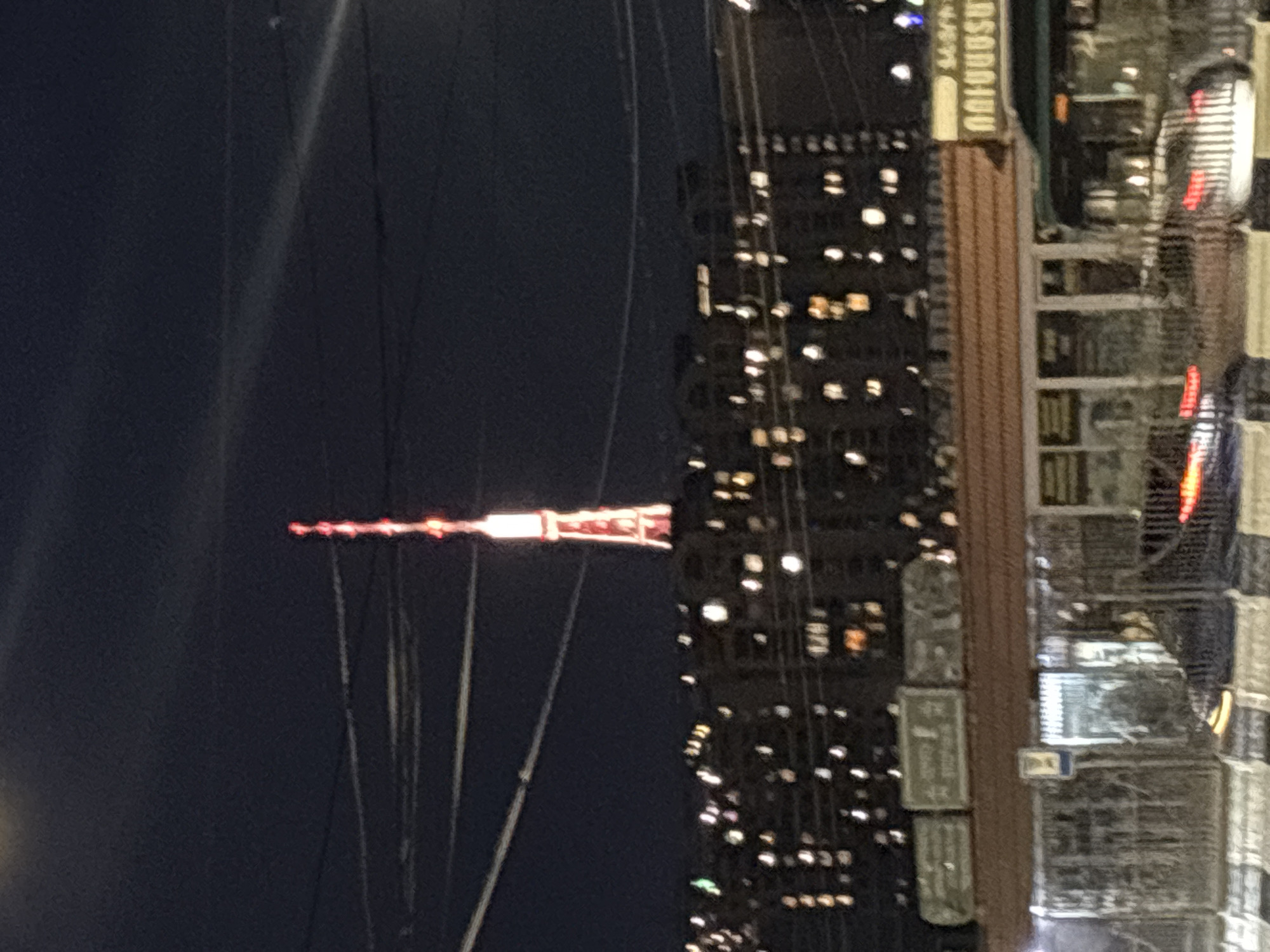 This image captures an urban night scene, likely in Yerevan, Armenia. The primary subject is a tall, slender, illuminated structure, strongly resembling the Yerevan TV Tower, standing prominently against a dark night sky. The top section of this tower is adorned with multiple red lights, while a substantial portion below glows brightly with white or yellow light. Several thin wires or cables are visible extending from the tower, arching across the dark sky.
To the right of the tower, a multi-story building dominates the right side of the frame. Numerous windows on this building are lit, casting a warm, yellowish glow onto the dark facade. On the upper part of this building, a light-colored sign is visible, displaying text in Armenian script. The clearest part of the text reads "ԱՆՈՒՇ" (Anush), a common Armenian name and word for "sweet," suggesting a potential business like a confectionery or a shop named after someone. Below this sign, other architectural details and dimly lit sections of the building are present.
The photograph depicts a static night landscape with no discernible human activity. The overall impression is one of urban quietude after dark, illuminated by artificial lights. The presence of the distinctive tower and Armenian script on the building sign strongly hints at the specified location of Yerevan, Armenia. The image itself appears to be rotated 90 degrees clockwise from its natural upright orientation.