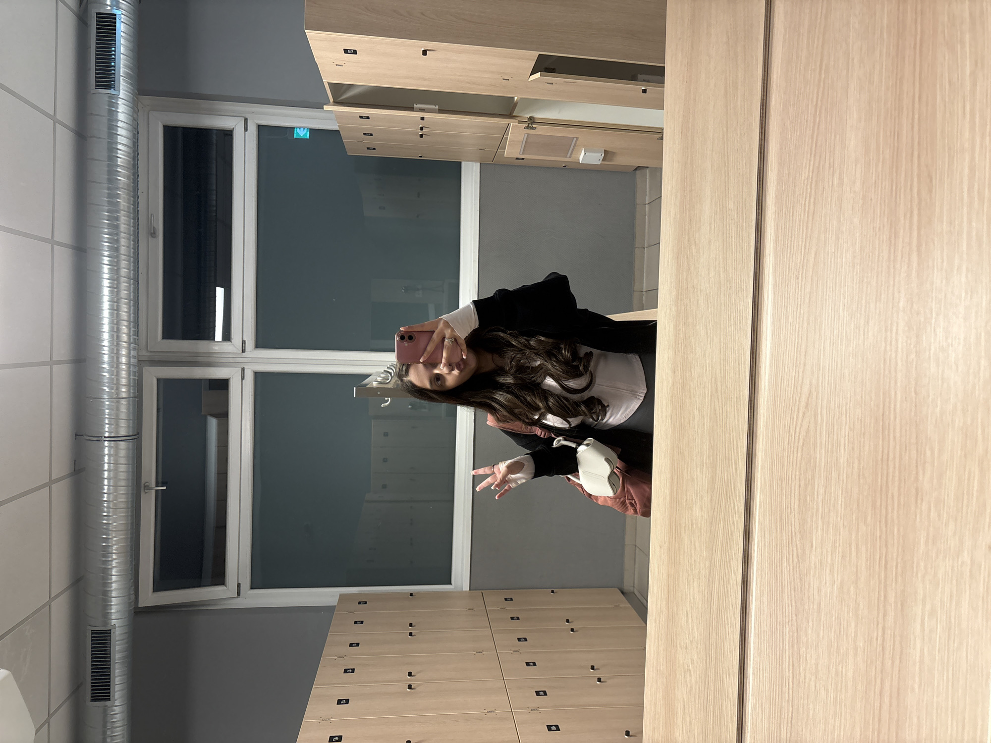 A medium, indoor shot depicts a woman posing for a selfie, likely in a changing room or locker room. The room is lined with light-colored wooden lockers, arranged in rows. Behind the woman, a window provides natural light. A metallic ventilation pipe runs vertically along the wall to the left of the window.
The woman holds a pink smartphone and makes a peace sign with her fingers. She has long, dark, wavy hair and wears a white top underneath a black jacket. She also carries a small white purse with a pink strap.
While the exact location in Mönchengladbach, Germany, cannot be determined from the image alone, the presence of lockers and the general appearance suggest a gym, sports facility, or public swimming pool changing room.