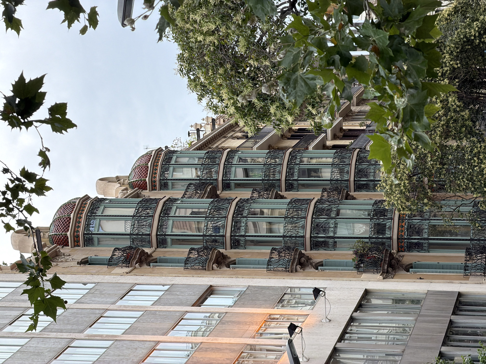 The image shows a row of buildings in Barcelona, Spain. The building on the left is a modern, gray concrete structure with many rectangular windows. The building to its right is older, with a beige facade and ornate green bay windows with black wrought-iron balconies. The bay windows are decorated with colorful tiled roofs. Trees with green leaves and small white flowers are visible in front of the buildings, obscuring some of the lower floors. The sky is overcast and cloudy. There are no people visible in the image. The scene appears to be a street view in a city.