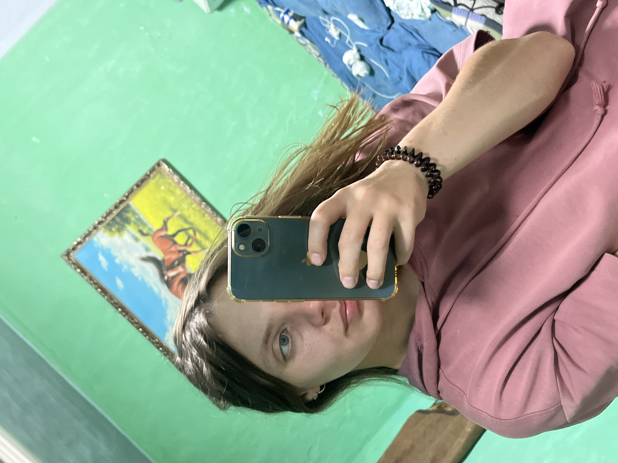 The image shows a young woman taking a selfie in a room with bright green walls. She is wearing a pink hooded sweatshirt and a dark coiled bracelet on her left wrist. She is holding a dark-colored smartphone in her right hand, with the camera facing her. A framed picture of two horses running in a field is visible on the wall behind her. A blue bed with white string-like objects is also present in the background. The lighting is somewhat dim, casting shadows on the wall. The overall impression is that of a casual, personal moment captured in a private space. There is no indication of the location being in Nizhny Tagil, Russia, or any specific city.